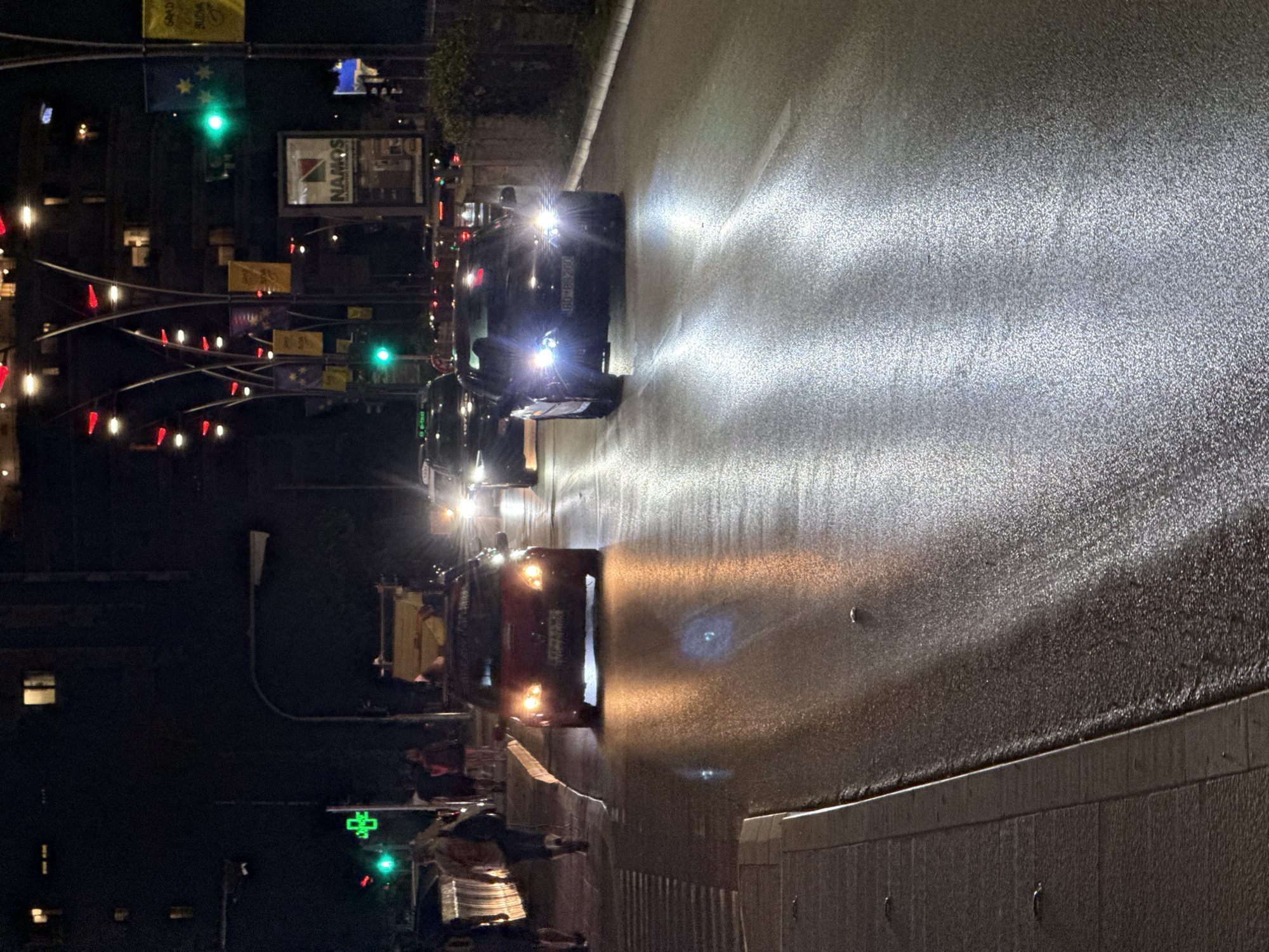 The image captures a nighttime scene on a wet street in Budva, Montenegro. In the foreground, the wet asphalt reflects the lights of passing vehicles and streetlights, creating streaks of illumination. A white pedestrian crossing is visible on the left side of the road, with a paved sidewalk adjacent to it.
Several vehicles are present on the road. A red car, facing the viewer, is in the left lane, its headlights illuminated. To the right of the red car, a black car, also facing the viewer, is in the right lane with its headlights on. Further down the road, more vehicles are visible, their lights suggesting ongoing traffic.
On the left side of the street, a few pedestrians are walking, silhouetted against the background. One individual appears to be carrying a backpack. Above them, a green cross symbol is illuminated, indicating a pharmacy. Traffic lights are also visible, with one showing a green light.
The background consists of buildings with illuminated windows and decorative streetlights with red and yellow accents. Banners with yellow and blue designs, possibly national flags or event decorations, are hung on poles along the street. A billboard with the "Namos" logo is also present. The overall atmosphere is that of a busy urban street at night, with the wet road adding a distinct visual element.