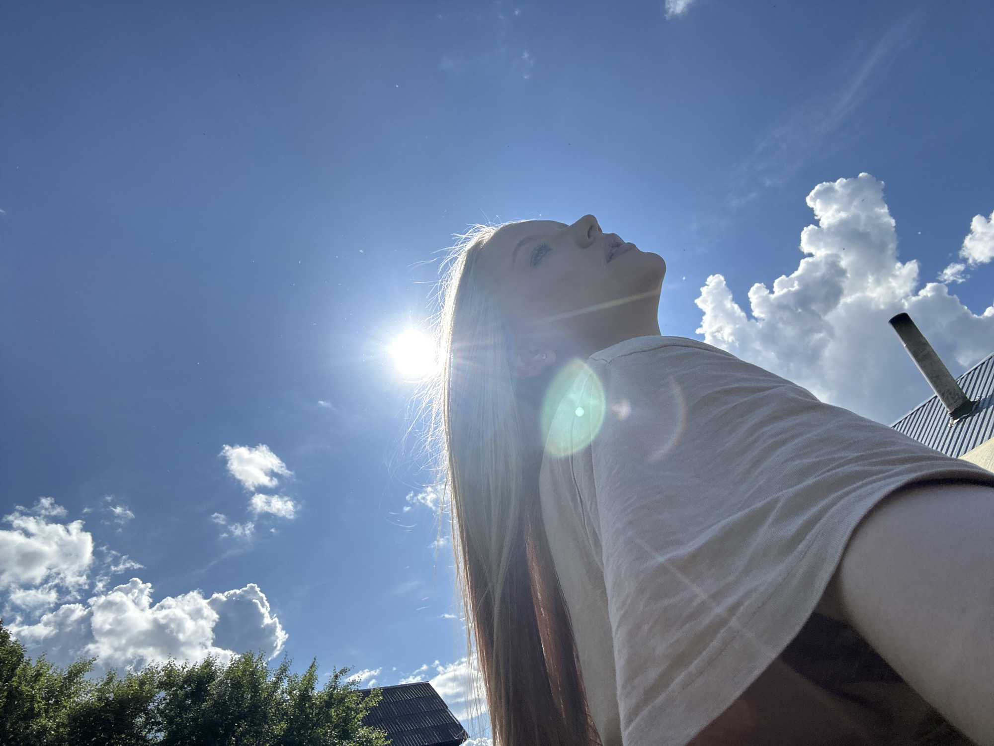 A low-angle shot captures a person looking up at a bright, sunny sky in Chaikovsky, Russia. The individual has long, light-colored hair and is wearing a light-colored shirt. The sun is shining brightly, causing lens flare. Scattered clouds are visible in the blue sky. Trees and buildings are partially visible in the lower portion of the frame. A metal pipe extends from the roof of a building.