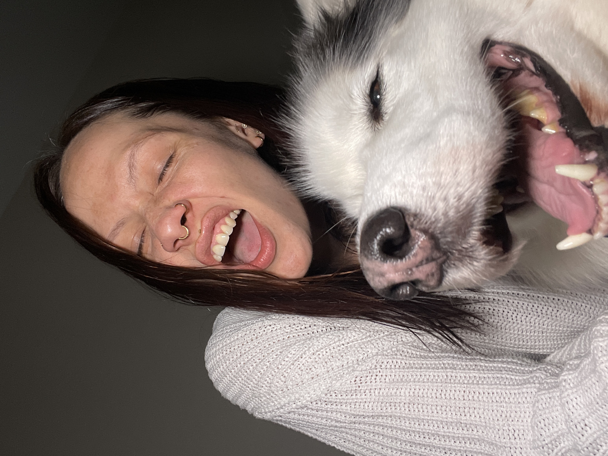 A woman and a white dog are closely positioned in a selfie-style photograph. The woman has dark hair, fair skin, and is wearing a gold nose ring. She has her eyes closed and mouth open in a wide smile or grimace. Next to her face is the head of a large white dog with black markings. The dog's mouth is also open, revealing its teeth and tongue. Both the woman and the dog are wearing a white knit sweater. The background is a dark grey. The photograph is rotated slightly counter-clockwise. There is no indication of Izhevsk, Russia, in this image.
