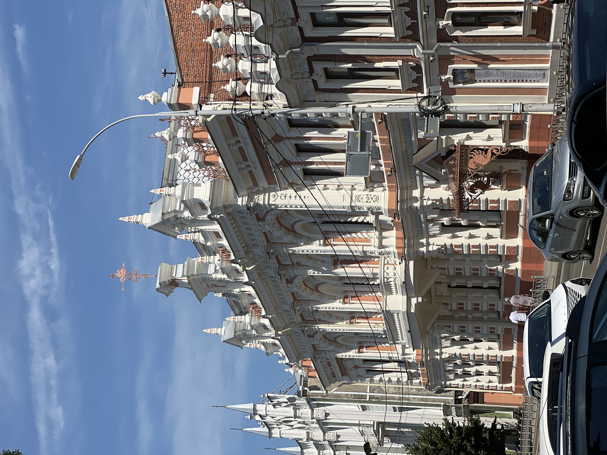 The image shows a street scene in Krasnodar, Russia, featuring ornate architecture and urban elements. In the foreground, several cars are parked or driving on the road. A white SUV is prominently visible, followed by a silver sedan. Further back, a white sedan with a distinct grille is also present.
The middle ground is dominated by a multi-story building with elaborate decorative details. The facade is painted in white and a pale salmon color, with intricate plasterwork, columns, arched windows, and balconies. A weather vane with a figure is perched on the rooftop. To the left of this building, another structure with pointed spires is visible, exhibiting a different architectural style.
In the background, the sky is a clear blue with scattered white clouds. A street lamp with a modern design arches over the scene. Two individuals, a man and a woman, are walking away from the camera on a sidewalk in front of the ornate building. The environment appears to be a sunny day in an urban setting, likely a historical district given the architectural style.