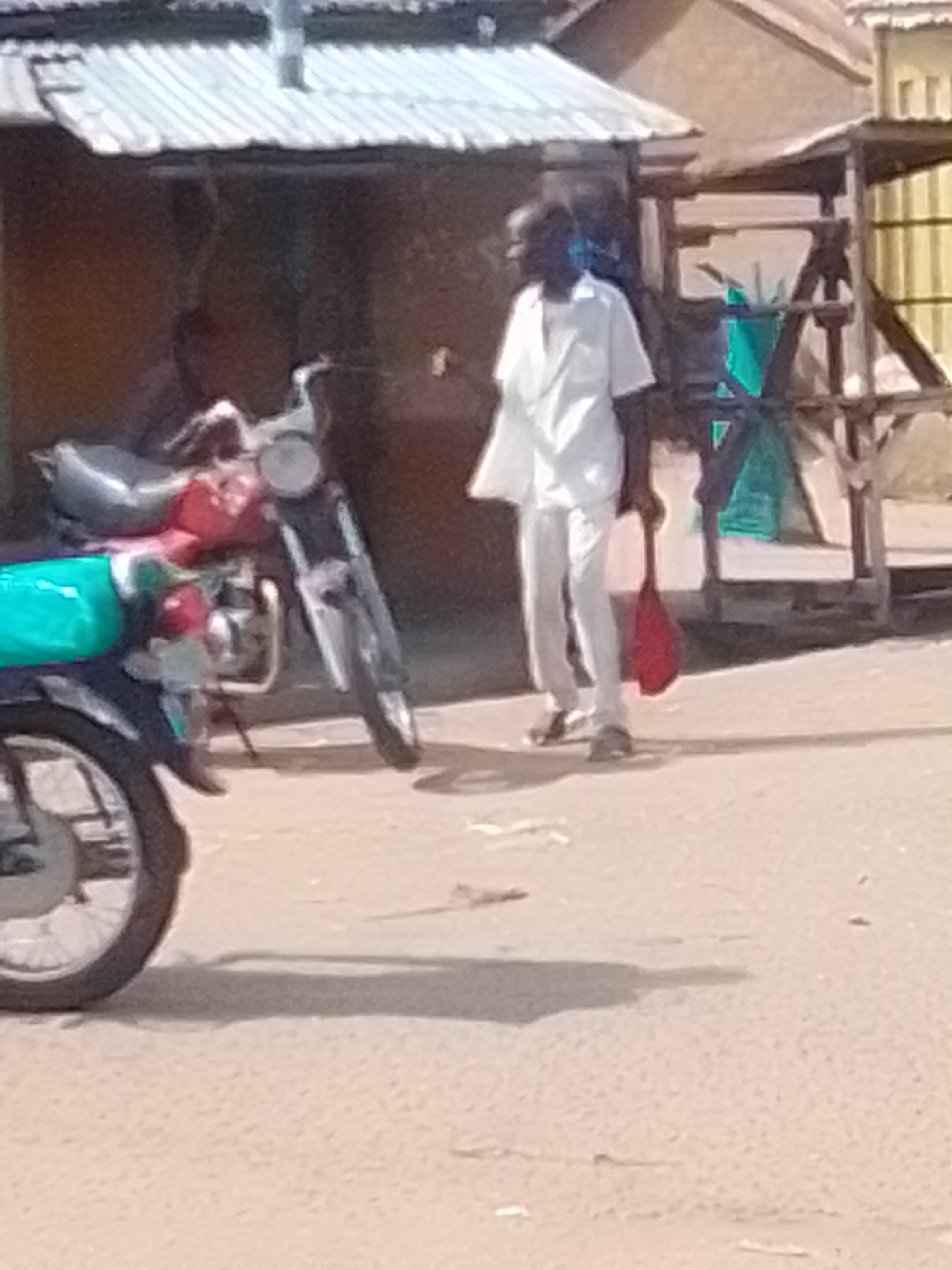 A man wearing a white shirt and light-colored trousers stands in an outdoor setting, holding a red bag. He is positioned in front of a motorcycle which is partially visible on the left side of the image. The motorcycle has a red fuel tank and a black front fender and wheel. Behind the man, there appears to be a small structure with a corrugated metal roof and wooden supports. The ground is a dirt or sandy surface. The background includes a building with a yellowish facade and more wooden structures. The scene is likely in Talasse, Nigeria, given the context provided.