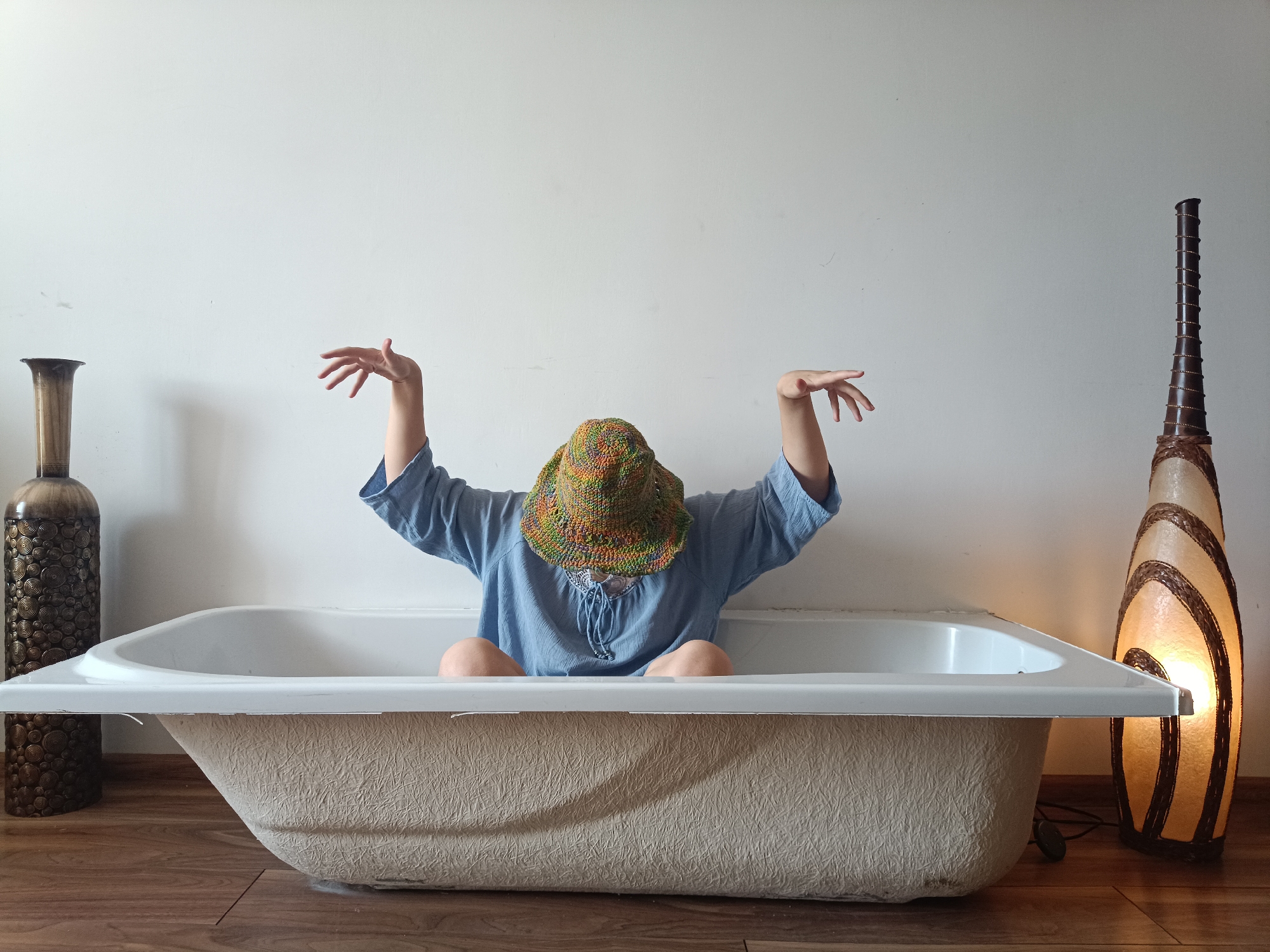 A person wearing a blue shirt and a colorful crocheted hat is sitting in a white bathtub. Their arms are raised outwards and upwards, with their hands open. The bathtub is placed on a wooden floor in front of a plain white wall. To the left of the bathtub is a tall, dark brown vase with a patterned surface. To the right of the bathtub is a tall, cream-colored lamp with dark brown woven accents, which is lit from within, casting a warm glow. The scene is indoors, with no specific indications of its location in Łódź, Poland.