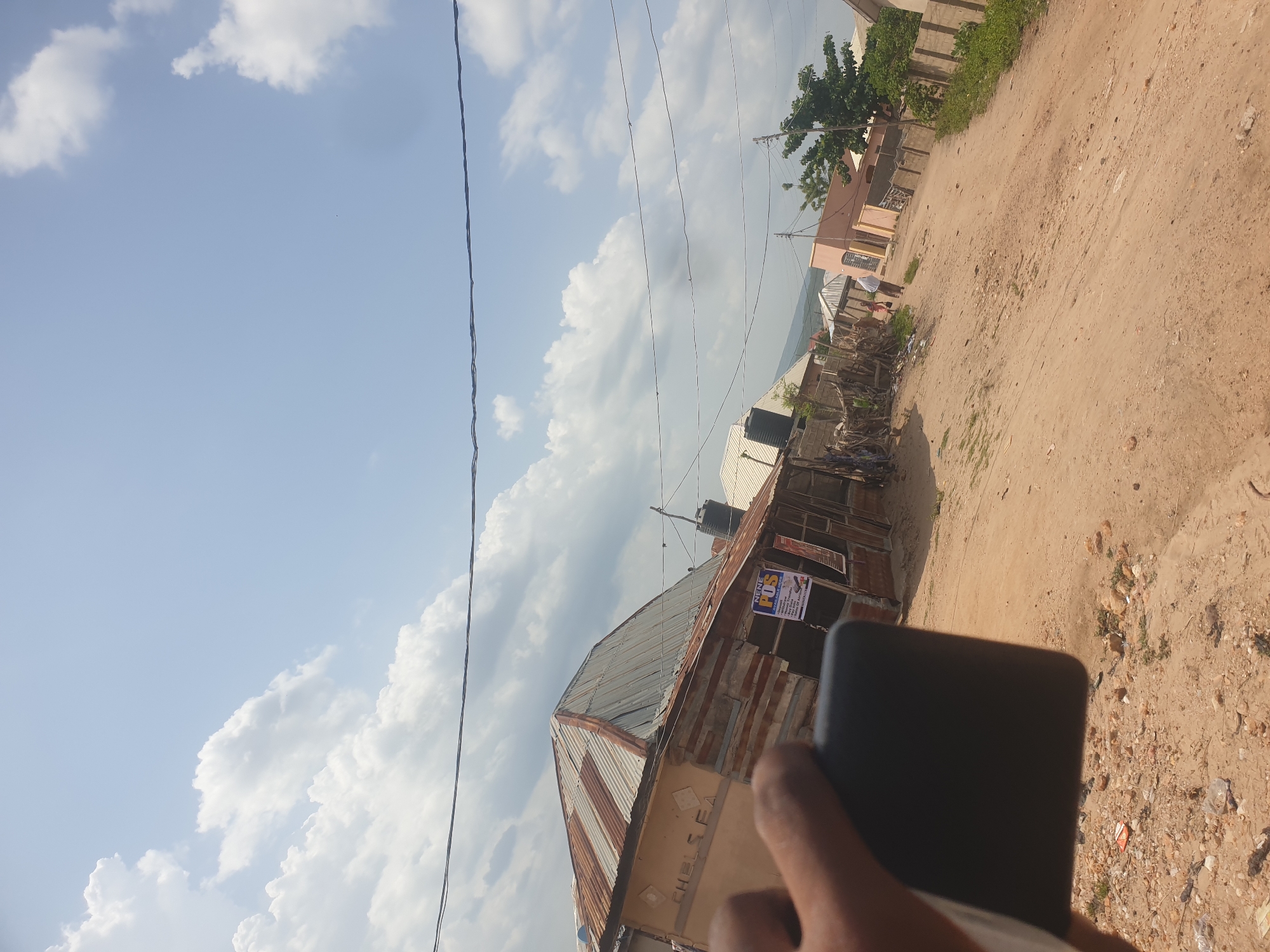 The image, taken in Zango-ndaji, Nigeria, shows a dirt road lined with buildings. The buildings are made of corrugated metal and cinder blocks, and some have water tanks on their roofs. Power lines are strung across the sky. The sky is blue with white clouds. A black object is partially visible in the foreground, possibly a phone held by a person. There is trash on the ground near the road. One building has a sign with "NENE POS" written on it. The picture is taken at an angle.