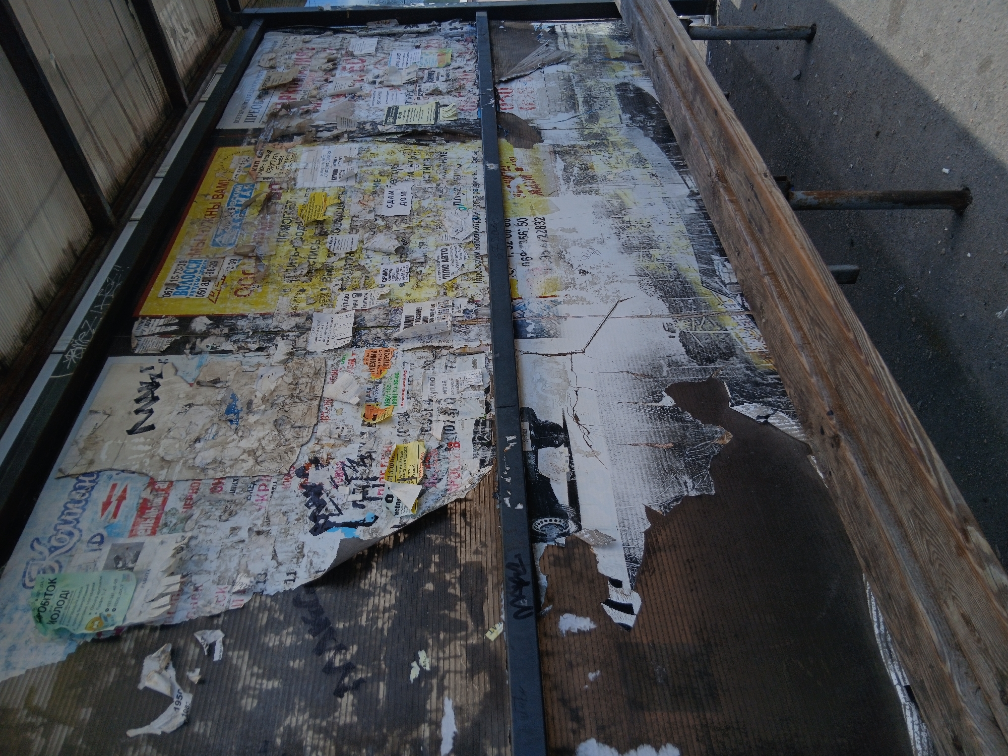The image depicts a weathered bus stop in Dnipro, Ukraine. The back wall of the bus stop is covered with numerous torn and layered advertisements, posters, and graffiti. The posters are a mix of colors, predominantly white and yellow, with some featuring red and blue text. Some posters have been partially peeled off, revealing the corrugated plastic underneath. The wall also shows signs of wear and tear, with streaks of dirt and discoloration.
In front of the wall, a wooden bench with metal legs is positioned. The bench appears old and worn, with visible wood grain and some damage. The ground in front of the bench is paved with asphalt, with small pebbles and debris scattered across it. The overall scene suggests a public space that has seen significant use and exposure to the elements. The lighting indicates it is daytime, casting shadows from the bus stop structure onto the ground. There are no people visible in the image.