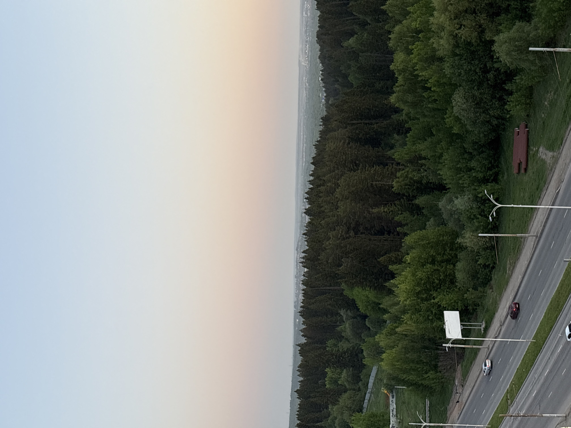 The image shows an elevated view of Izhevsk, Russia. The sky transitions from light blue-grey to a soft orange hue, indicating either sunrise or sunset. A dense forest borders the city, its trees a mix of dark green. A roadway with multiple lanes runs parallel to the forest edge, featuring streetlights and several cars traveling on it. A white billboard is positioned alongside the road. A section of red-brown roofing material lies on the grassy area near the forest. The image has been rotated 90 degrees counterclockwise.