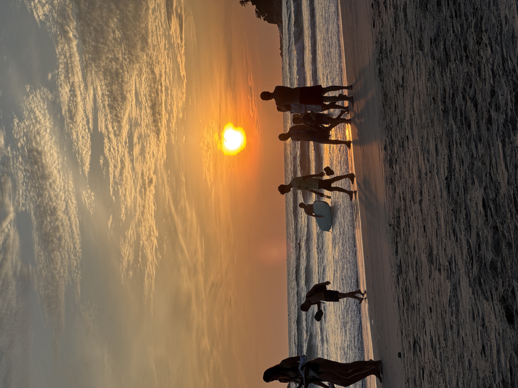The image captures a group of people walking along the beach in Karon, Thailand, during sunset. The sun, a bright orange orb, is low in the sky, casting a warm glow and long shadows on the sand. The sky is filled with streaks of orange, yellow, and white clouds, creating a dramatic backdrop. The ocean waves gently roll onto the shore, reflecting the sunset's colors. Silhouettes of palm trees are visible on a distant headland.
Several individuals are depicted in various states of movement. In the foreground, a woman in a bikini walks towards the left, carrying a towel. Beside her, a young boy walks away from the camera, looking down. Further along the beach, a group of people are walking together. One person is carrying a surfboard, while others appear to be strolling and talking. The general atmosphere is one of leisure and enjoyment of the coastal scenery at dusk.