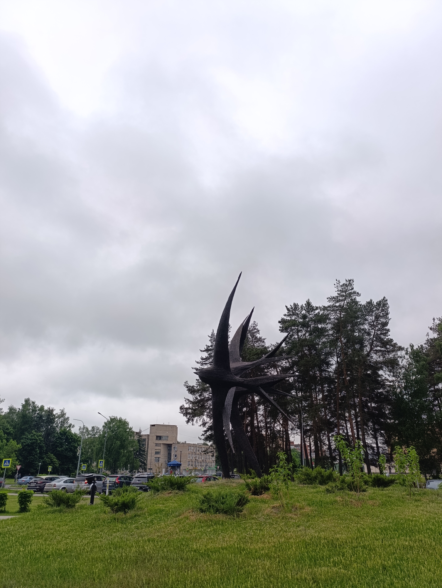 The image shows a park with a modern sculpture in the city of Unknown, Unknown. The sculpture, dark in color, has several pointed extensions reaching upwards. Behind the sculpture are tall pine trees. In the background, there is a multistory building and a street with parked cars. The sky is overcast, and the foreground consists of a grassy area with some shrubs.