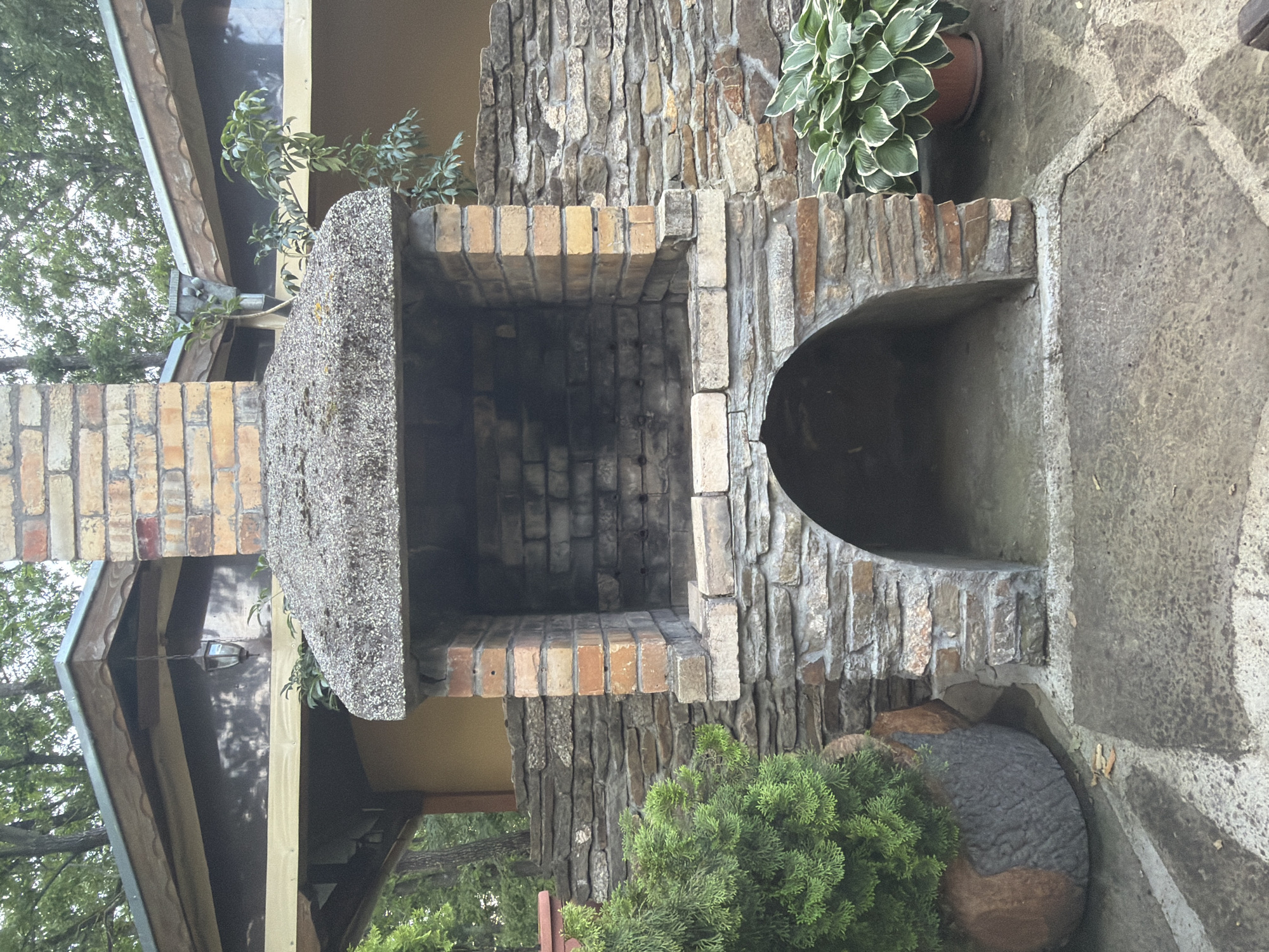 The image captures an outdoor barbecue and fireplace structure crafted from a combination of brick, stone, and concrete. The central feature is a rectangular firebox constructed from multi-toned bricks, showing signs of use with darkened interior walls. A thick, grey concrete slab forms the roof over the firebox, extending upwards as a chimney base. To the right, an arched opening is integrated into a rough-hewn stone and brick wall, providing a niche or secondary cooking area.
Green foliage surrounds the structure. A potted Hosta plant with variegated leaves is placed on a stone shelf to the right of the arched opening. Below, a dense green conifer or shrub thrives near the base, alongside a textured, spherical garden ornament in grey and brown. In the background, elements of a building are visible, including wooden eaves, a dark reflective surface (possibly a window or panel) with a reflected outdoor light fixture, and a portion of a roof. Mature trees with lush green leaves are seen beyond the building.
The ground surface consists of irregularly shaped grey flagstones, forming a patio area. The lighting suggests it is daytime, with soft, diffused light. There are no individuals present, and no specific activities are taking place. The scene depicts a serene and well-established outdoor space, set in Sushina, Bulgaria. No visible text is present in the image.