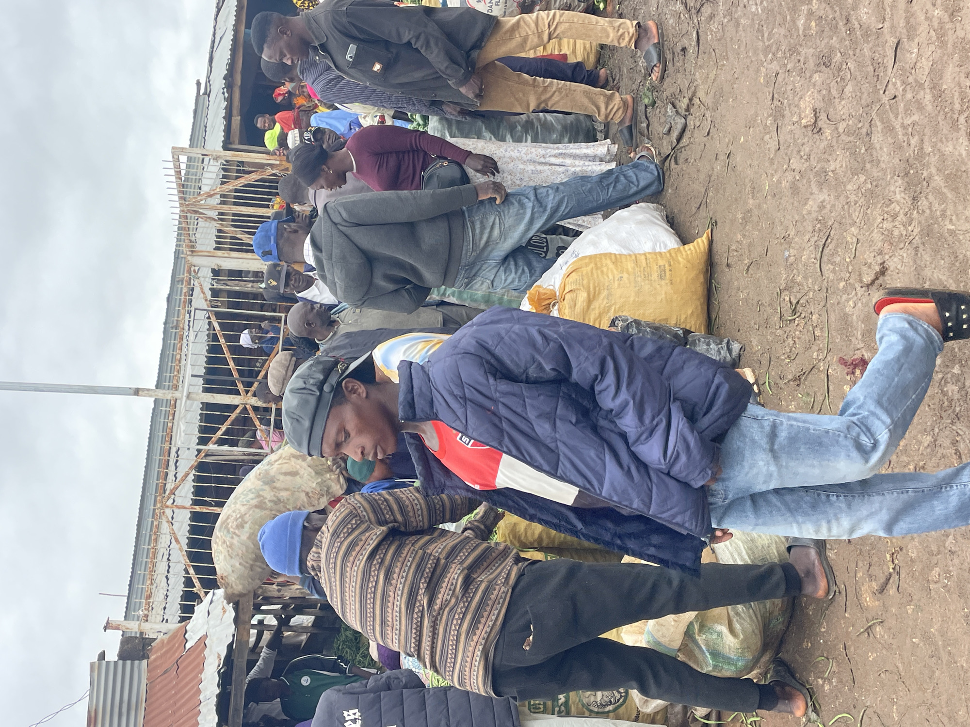 This image depicts a busy outdoor market scene in Gura Top, Nigeria. Several individuals are present, some interacting, while others appear to be shoppers or vendors. The ground is muddy, suggesting recent rain or a generally damp environment. Various sacks, some appearing to contain produce or goods, are visible throughout the scene, indicating commerce is actively taking place. The background shows rudimentary market stalls and structures, typical of an open-air market. The atmosphere is one of bustling activity.