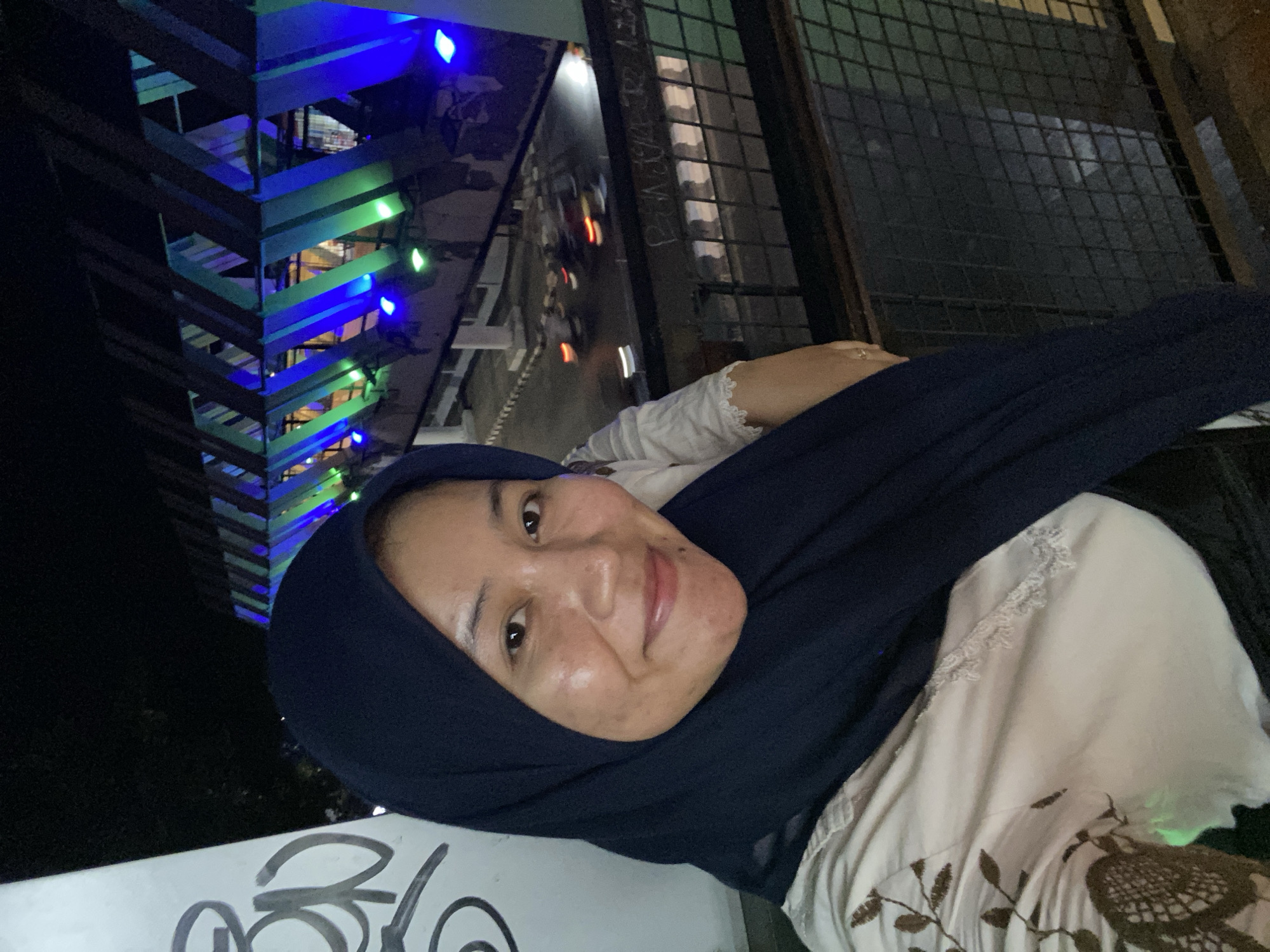 A woman wearing a dark blue hijab and a light-colored, patterned shirt poses for a selfie at night. She is standing on an elevated walkway or bridge with a metal railing and a grid pattern in front of her. In the background, a bridge structure is illuminated with blue and green lights, and traffic with blurred lights is visible on the road below. The scene appears to be in Banjarbaru, Indonesia, likely a public area with pedestrian access and decorative lighting. There is graffiti on a white pillar to the left of the woman.