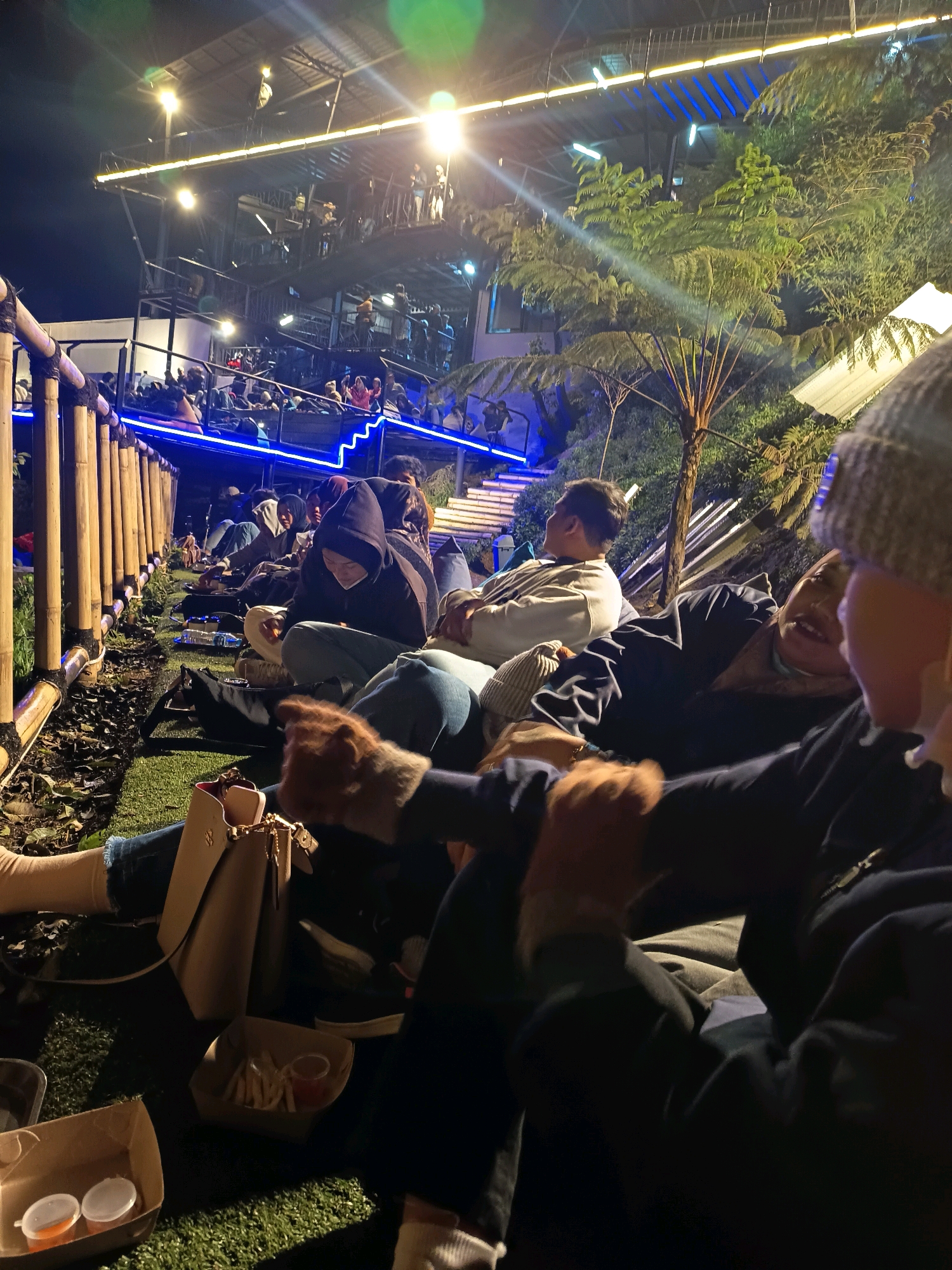The image depicts a nighttime scene at an outdoor venue, likely a restaurant or entertainment space, in Keditan, Indonesia. A group of people are casually seated on artificial grass, facing a multi-level structure that is brightly lit with yellow and blue lights. The structure appears to be a building with balconies or walkways, populated by more individuals. Lush green foliage, including ferns and trees, surrounds the area. In the foreground, several individuals are visible, some in focus and others blurred. One person in the immediate foreground wears a knitted beanie. In front of the seated individuals, there are food containers, including one with french fries and dipping sauce, and another with small containers of sauce or food. A beige handbag rests on the grass. The overall atmosphere suggests a relaxed social gathering during the evening.