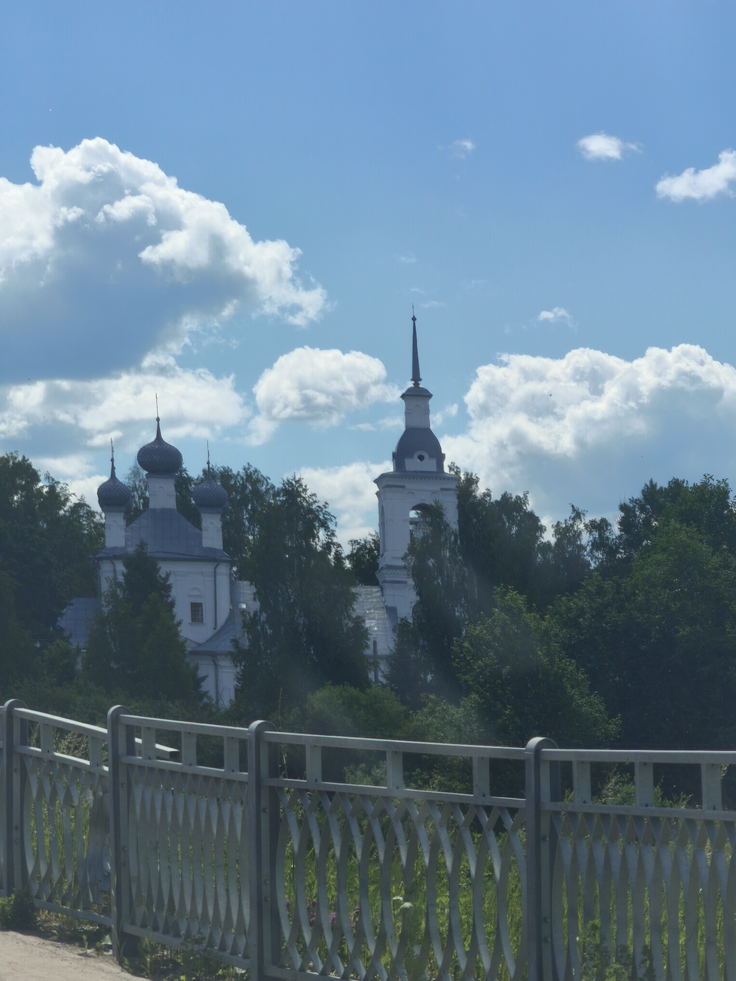 The image shows a view of a white church with multiple domes and a bell tower, surrounded by lush green trees. A decorative metal fence runs along the bottom of the frame. The sky above is blue with scattered white clouds. The church appears to be the Church of the Resurrection, located in Kady, Kostroma Oblast, Russia, as indicated by its architectural style and the presence of onion domes. The scene is a daytime outdoor view, likely taken from a slightly elevated position, possibly across a street or body of water, given the presence of the fence. There are no people or animals visible in the frame.