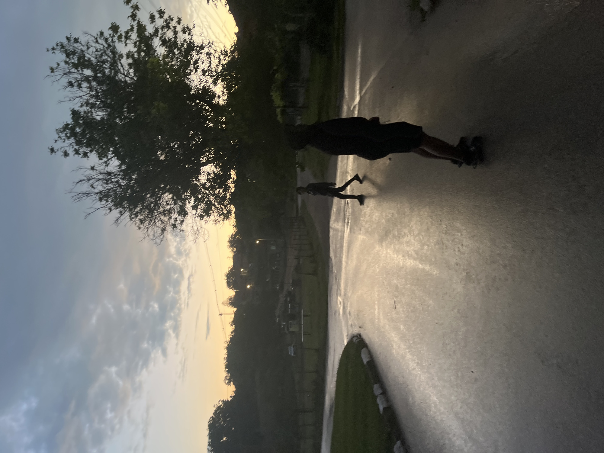 The image, taken in Bukovlak, Bulgaria, depicts an outdoor scene at dawn or dusk, characterized by a gradient sky transitioning from orange-yellow at the horizon to pale blue-grey overhead, with indistinct cloud formations. A wet, paved road occupies the foreground and mid-ground, exhibiting reflective streaks from ambient light. To the far left, a patch of green grass is bordered by a curb featuring black and white markings. Two silhouetted individuals are visible on the road. In the foreground right, a person is partially visible from the waist down, wearing dark shorts and sneakers, standing on a skateboard. In the mid-ground left, another person walks from left to right, clad in dark clothing. Dense trees and foliage frame the scene, with a prominent leafy tree on the mid-right side. In the distant background, obscured by vegetation, are indistinct structures, a partially visible white vehicle, and illuminated streetlights or utility poles. The wet road surface indicates recent precipitation.