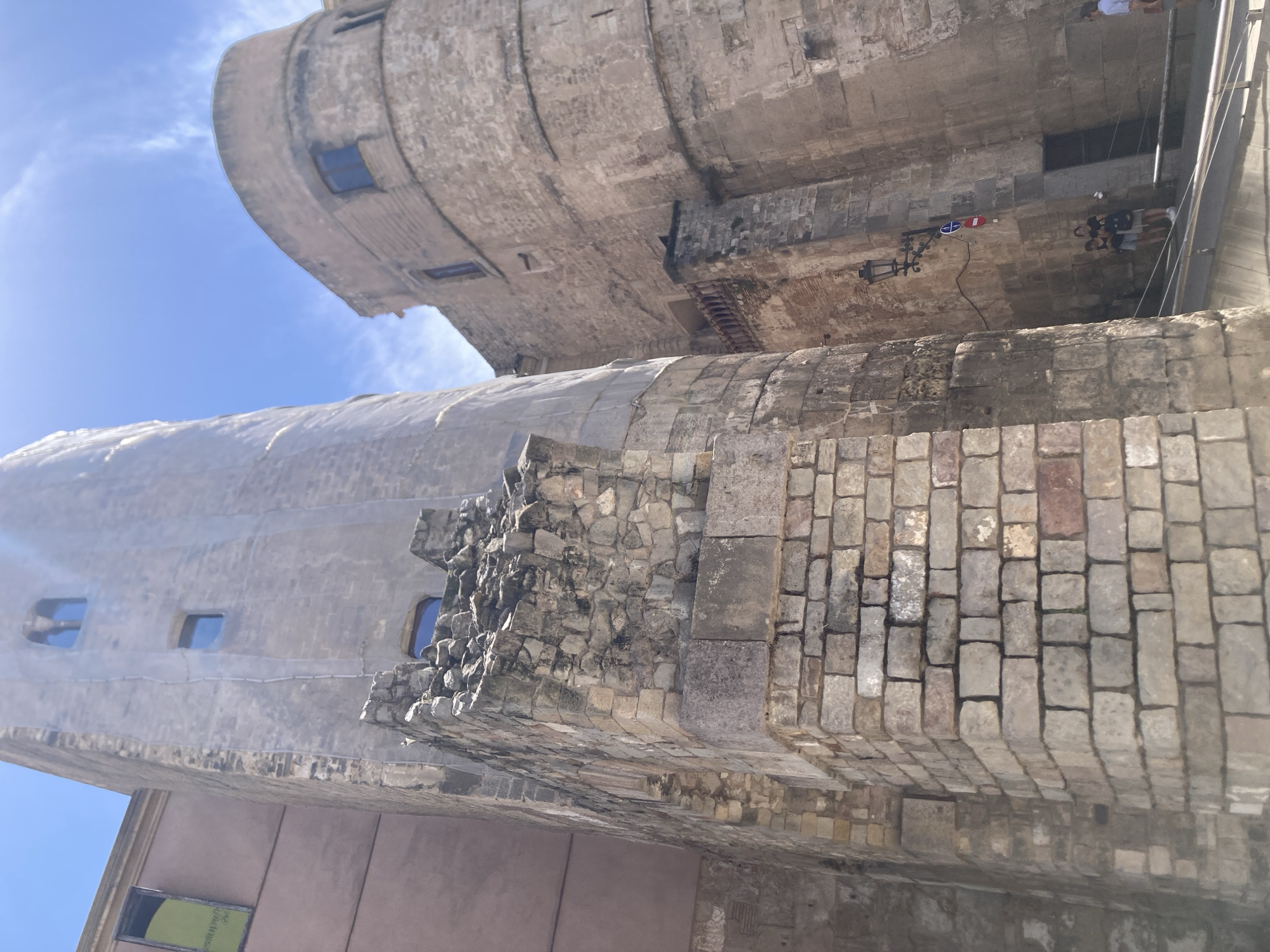 The media file captures a low-angle, upward view of a large, ancient stone structure, likely a tower or part of a fortification, in Barcelona, Spain. The stonework is varied, with some sections appearing weathered and rough, composed of irregular stones, while others are meticulously laid with rectangular blocks. The structure shows signs of age and possibly some restoration or ongoing work. Small, dark rectangular windows are visible on the curved surfaces of the tower.
The sky above is a clear, bright blue, suggesting a sunny day, likely mid-morning or early afternoon due to the quality of the light. Sunlight casts shadows and highlights the textures of the stone. In the distance, above the stone structure, hints of a modern building with glass windows can be seen.
Towards the right side of the image, several people are visible, appearing to be walking or standing near the structure. They are dressed in casual clothing, consistent with a tourist or visitor setting. One person is near a decorative, ornate lamp attached to the wall, which has a circular emblem with red and blue colors. The overall scene conveys a sense of historical grandeur and the present-day experience of exploring such a site. There is no discernible text visible in the image.