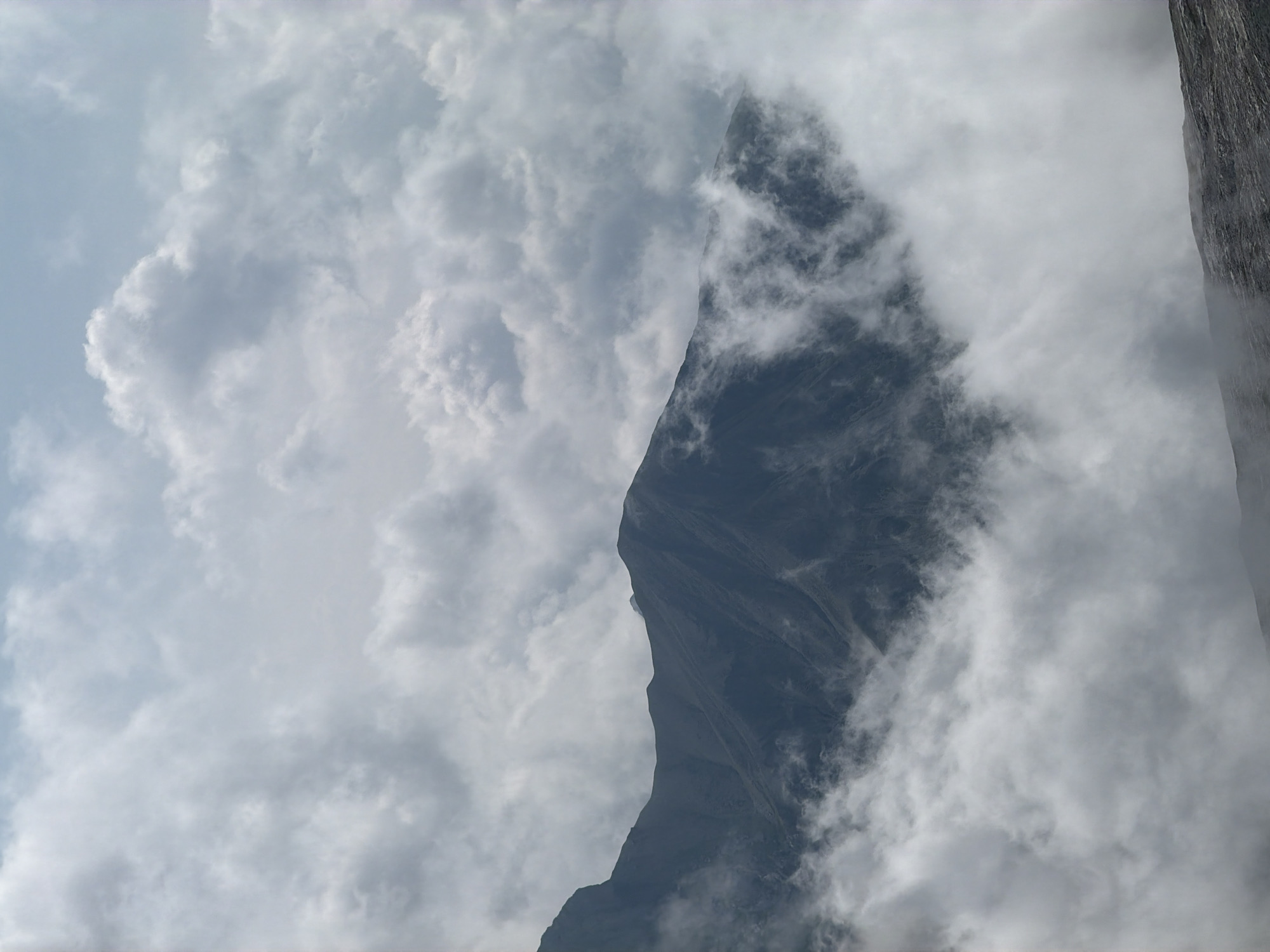 The image depicts a dramatic mountain landscape partially obscured by clouds. A dark, rugged mountain range dominates the mid-ground, with its peaks and slopes softened by the swirling mist. The sky above is filled with a dynamic pattern of light and dark clouds, suggesting a changeable weather system. The foreground shows a rocky, uneven surface, possibly part of the mountain's lower slopes or a viewpoint.
This scene is likely Mount Wank, a prominent peak near Garmisch-Partenkirchen, Germany. Mount Wank offers panoramic views and is a popular destination for hiking and cable car rides. The cloudy conditions suggest it might be early morning or late afternoon, or simply a day with unsettled weather typical of the Bavarian Alps. There are no people or man-made objects visible, emphasizing the natural grandeur of the mountainous environment.