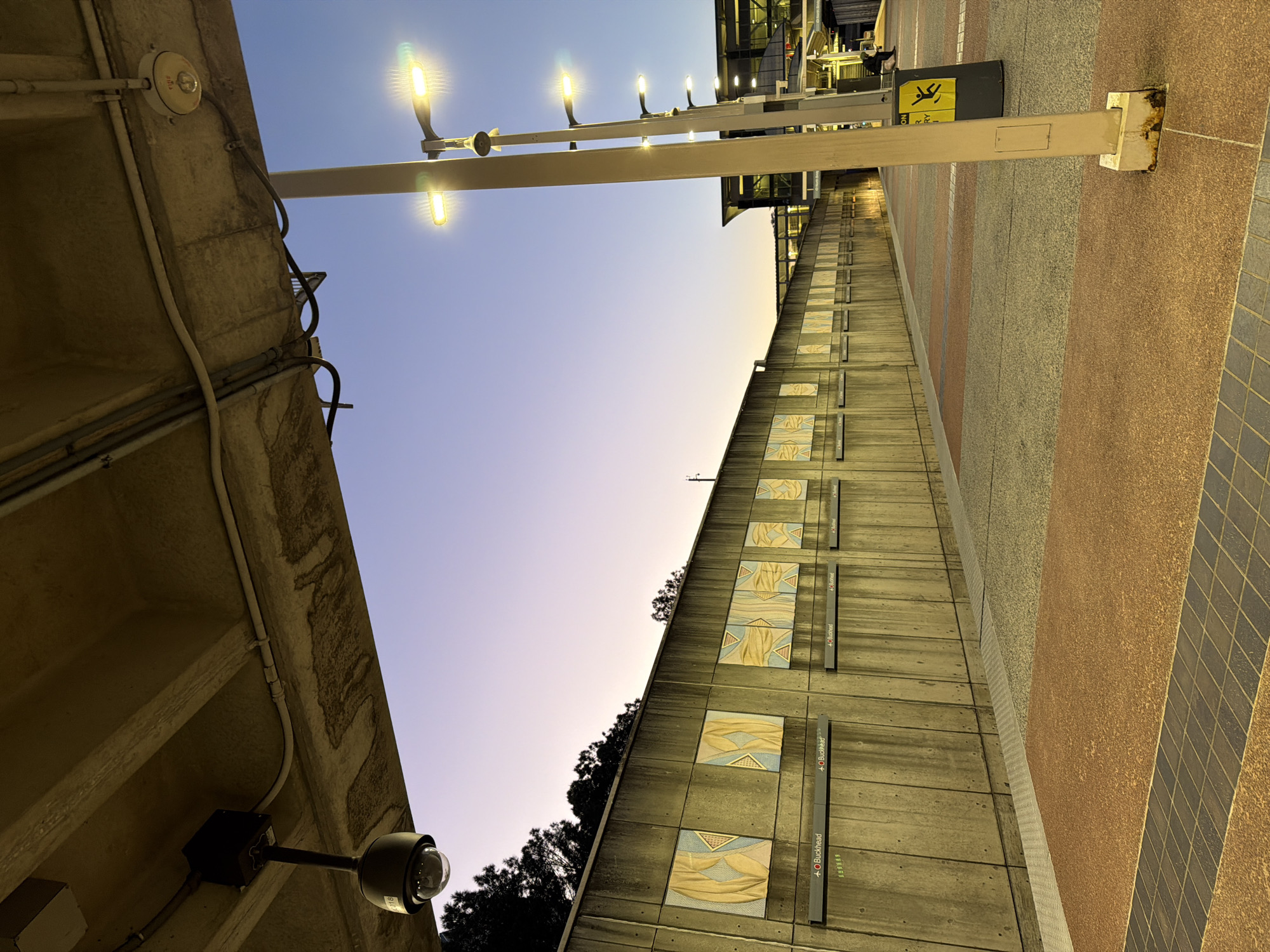 The image shows a portion of the Buckhead MARTA station in Atlanta, Georgia. The view is an angled upward shot of the station platform at dusk. A concrete wall with decorative tile panels and station signs (labeled "Buckhead") runs along the length of the platform. Above the wall, there is a line of trees and the evening sky transitioning from blue to orange. Streetlights illuminate the platform. On the left, there is a concrete ceiling with electrical conduits and a security camera. The platform surface is made up of different colored tile, and there are signs indicating train directions and platform safety information. The architecture suggests it's a public transportation area, specifically a train or subway platform.