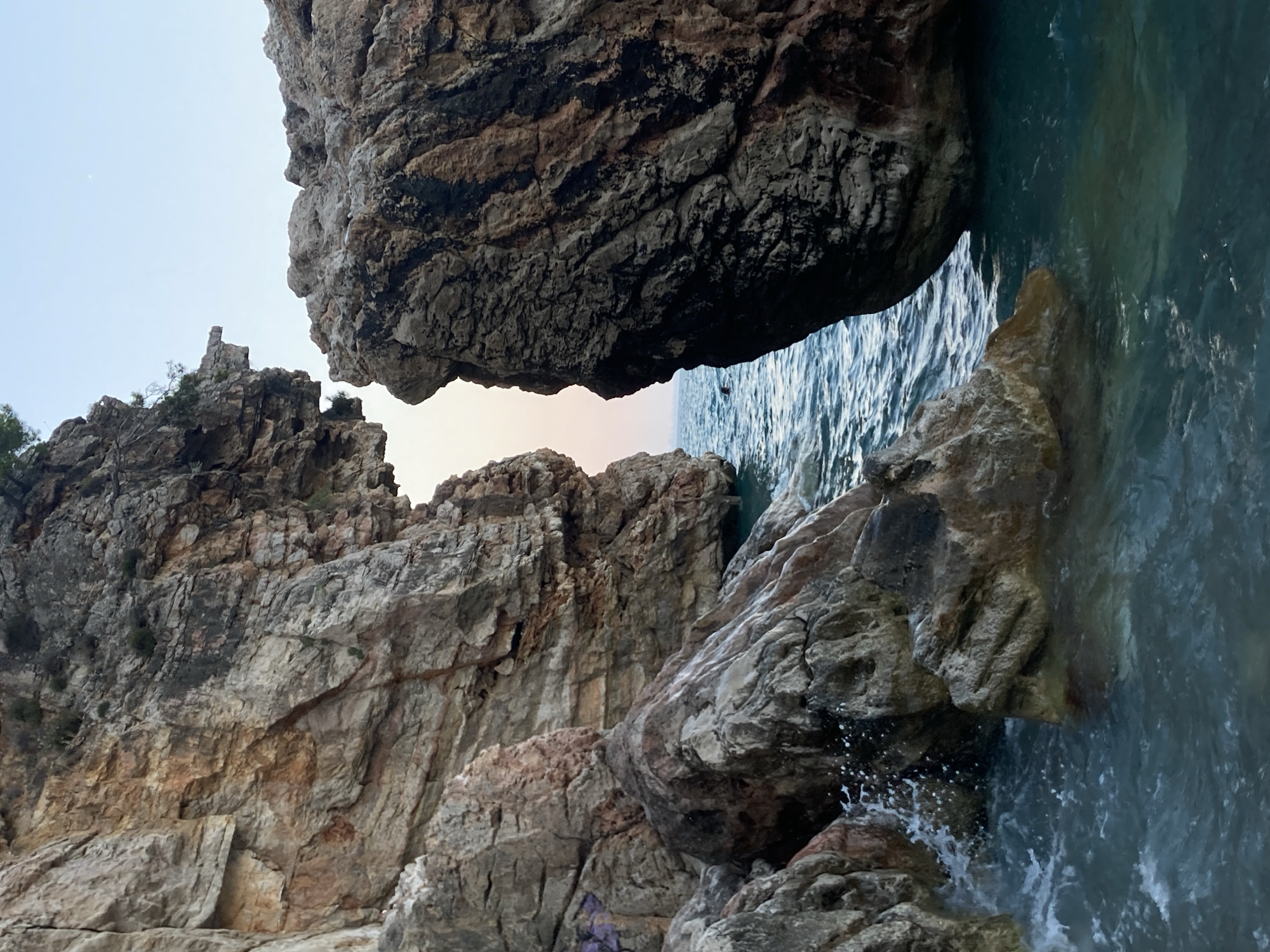 The image captures a dramatic view of the Mediterranean Sea crashing against rugged, weathered rock formations. The foreground is dominated by large, textured boulders and rocks, some of which are partially submerged in the churning, turquoise water. White spray is visible as waves break against the stones. In the mid-ground, a narrow opening between two massive cliffs reveals the horizon, where the clear sky transitions from a pale pink to a soft blue, indicating either sunrise or sunset. Two figures can be seen swimming in the water through this opening. The rock faces exhibit layers of sediment and some patches of vegetation clinging to the higher ledges. The overall scene suggests a natural, wild coastal environment. This scene is representative of the dramatic coastline found in and around Kemer, Turkey, known for its picturesque beaches and rocky coves.