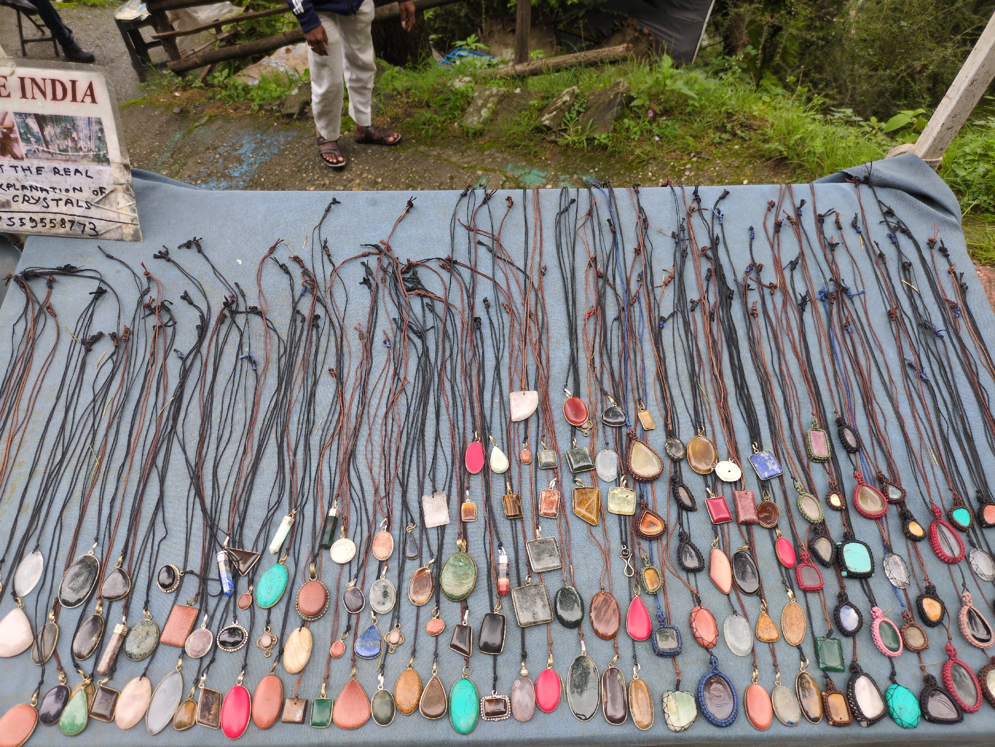 In Bhagsu, India, a stall displays a collection of necklaces with gemstone pendants. The necklaces are laid out on a blue fabric surface, with the strings hanging loosely. The pendants vary in shape, size, and color, featuring stones like turquoise, agate, quartz, and various other polished minerals. Some pendants are large and oval, while others are rectangular or in more abstract shapes. The setting appears to be outdoors, with greenery and a wooden structure visible in the background. A person's legs, wearing light-colored pants and sandals, are visible to the left of the stall. A sign with text, partially visible, is also present on the left side of the image.