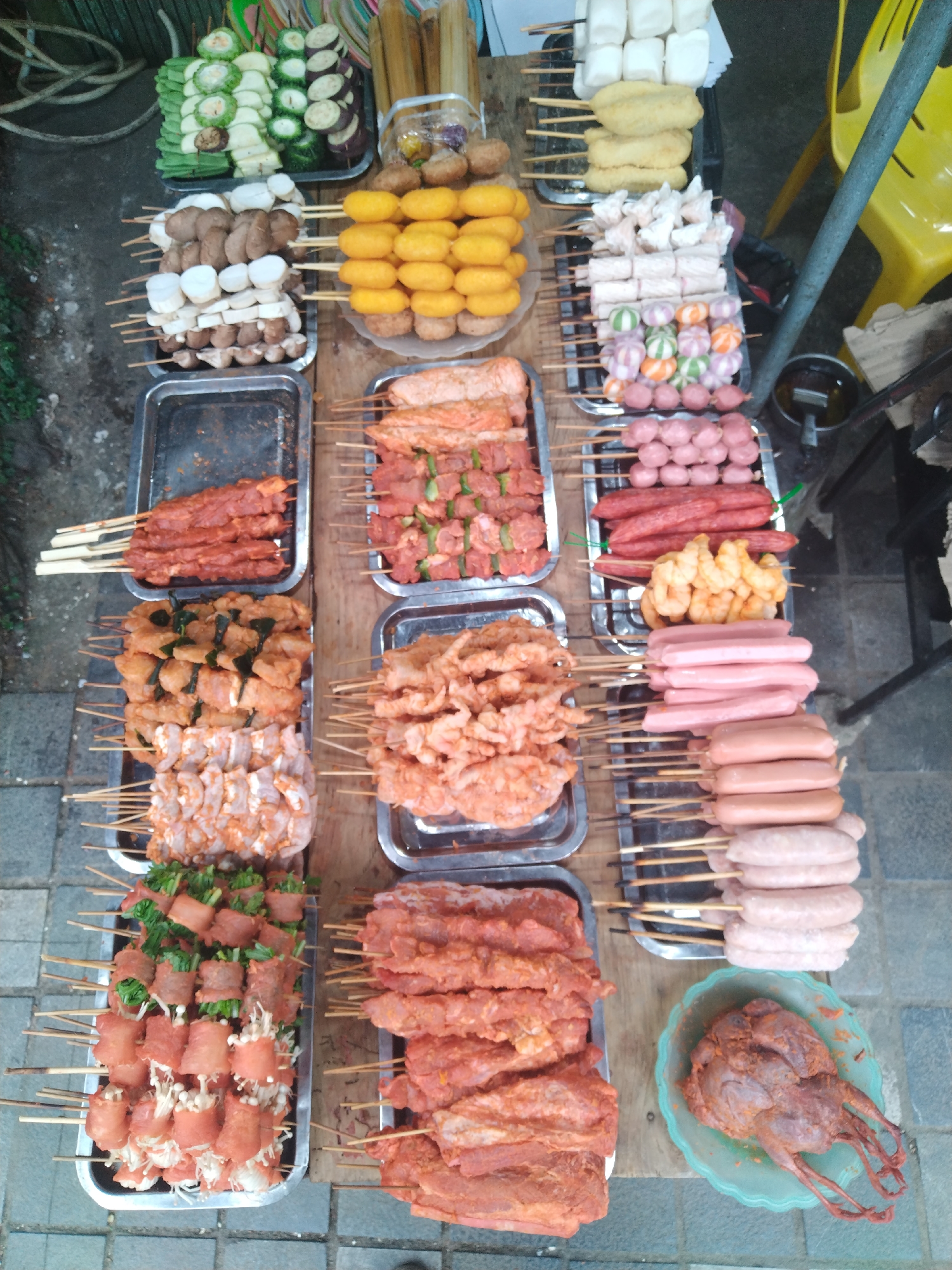 This overhead image showcases a vibrant street food stall, likely found in a market or roadside setting in Sa Pa Ward, Vietnam. The primary subjects are a diverse array of raw and marinated food items meticulously arranged on wooden and metal trays, primarily skewered and ready for cooking.
The stall features a wide selection of ingredients:
**Vegetables:** Skewers hold slices of green bitter melon, light green gourds (possibly zucchini or chayote), and purple eggplant.
**Mushrooms:** Both dark brown shiitake mushrooms and white cap mushrooms are visible on skewers.
**Processed Foods:** Various processed items include white tofu blocks, yellow fish cakes, an assortment of pink and reddish sausages of different sizes and shapes, and spherical fish or meat balls, some plain yellow and others striped green and orange.
**Meats and Seafood:** Numerous skewers are laden with marinated shrimp, raw chicken or pork pieces (some with dark green herbs/leaves), and red-marinated beef or pork chunks. There are also skewers featuring thinly sliced meat (possibly bacon) wrapped around enoki mushrooms or green onions.
**Other:** A distinct feature is a whole, marinated bird, possibly a quail or small pigeon, resting on a light blue plastic plate.
The scene depicts a typical setup for a street food vendor, where customers would select their preferred skewers for on-demand grilling or frying. The items are neatly organized, emphasizing variety and freshness.
Notable details include the outdoor setting, suggested by the paved ground and a partial view of a yellow plastic chair, a dark metal pole, and some green foliage in the background. The lighting suggests it is daytime. No people are visible, and there is no visible text. The overall atmosphere conveys the rich and accessible culinary experience common in Vietnamese street food culture.