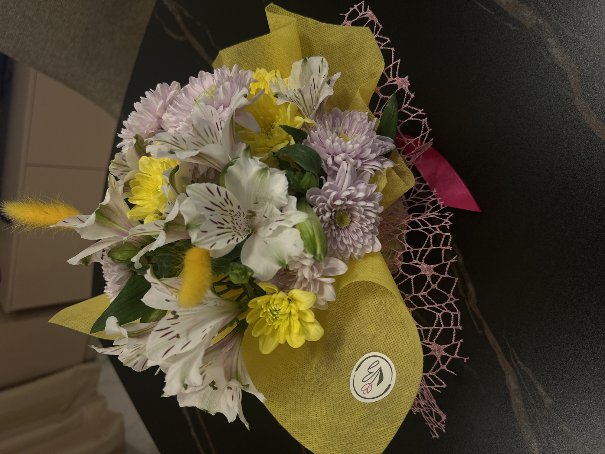 A close-up shot captures a bouquet of flowers presented on a dark, possibly marbled, surface. The arrangement includes white alstroemeria with maroon streaks, yellow and lavender chrysanthemums, and yellow lagurus ovatus. The bouquet is wrapped in yellow fabric, adorned with a sticker, and accented with pink net-like material and ribbon. The context of the scene lacks sufficient detail to definitively locate it within the city of Sofia, Bulgaria. The environment appears to be indoors.