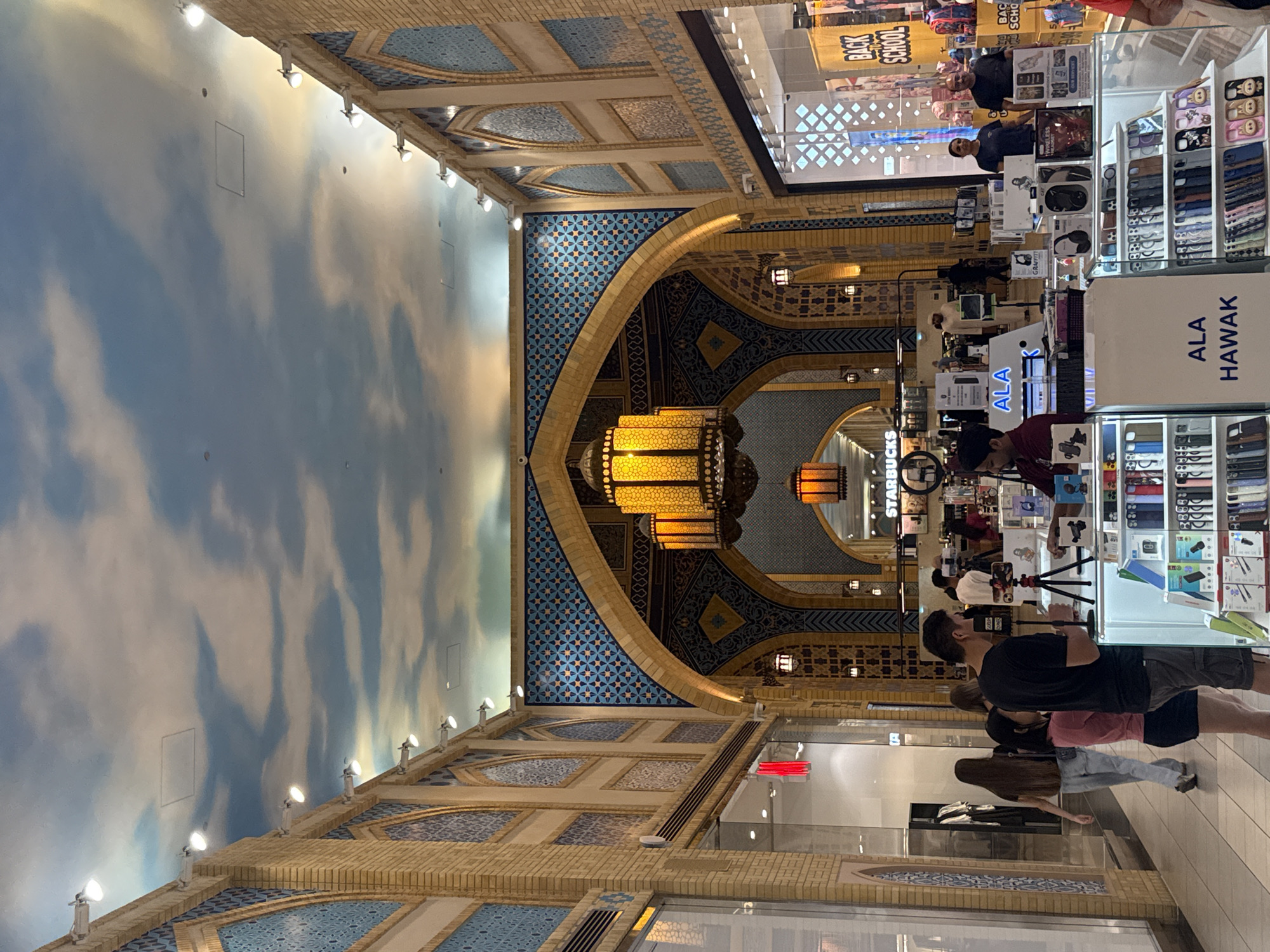 This is a photograph of the interior of a shopping mall in Dubai, United Arab Emirates. The architecture features Middle Eastern influences with arched doorways, patterned tilework in blue and gold, and decorative lanterns. The ceiling is painted to resemble a sky with clouds. In the foreground, there is a retail kiosk selling mobile phone accessories, with shelves displaying cases and other items. Two individuals are standing behind the kiosk, and a man is filming with a tripod in front of it. Further back, people are walking through the mall, and storefronts are visible on either side, including a "Starbucks" sign. The overall scene depicts a busy commercial environment within the mall.
