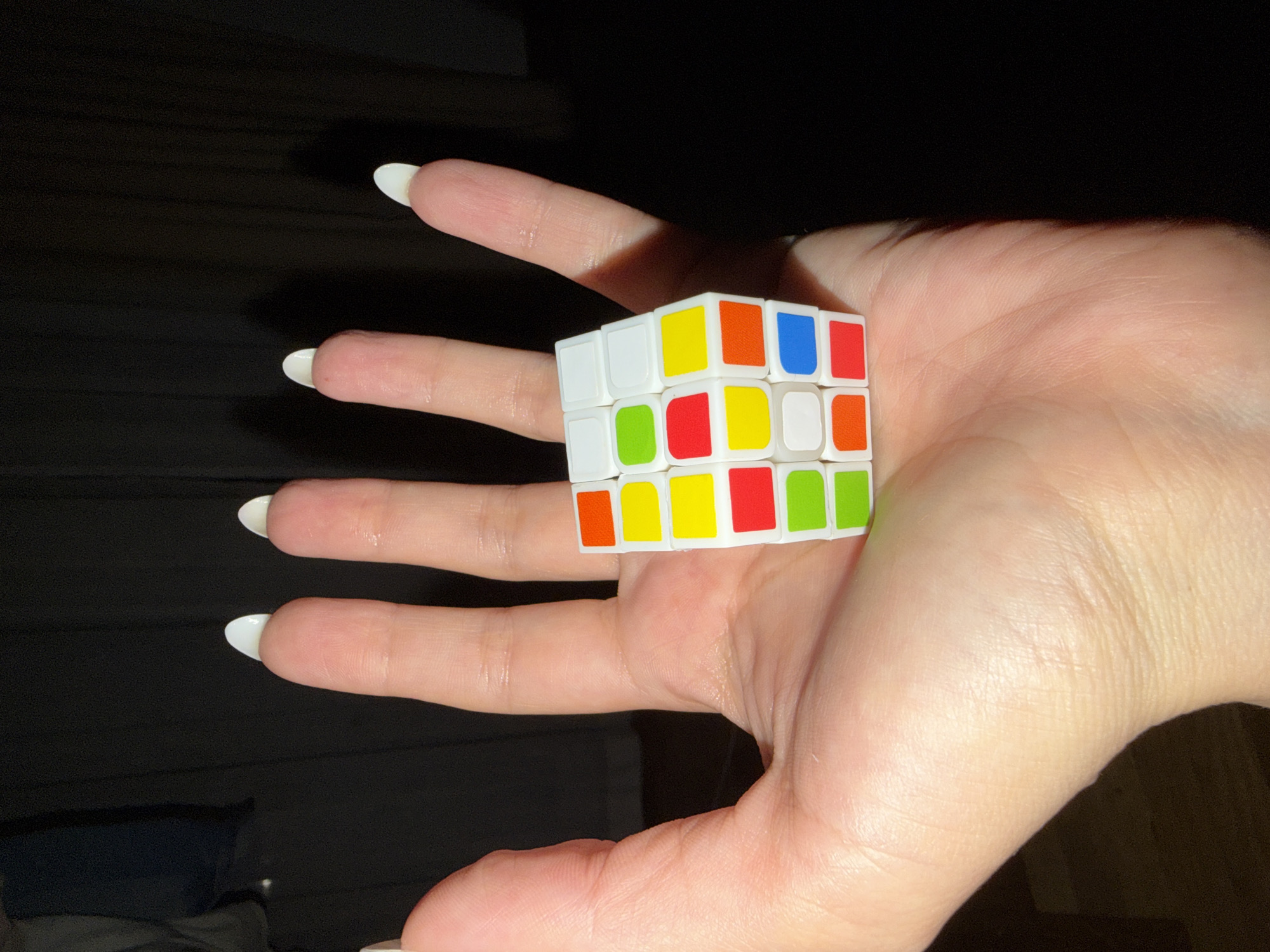 A hand holds a small, white Rubik's Cube. The Rubik's Cube features the standard colors: white, yellow, orange, blue, red, and green. The hand has long fingers with white polished nails. The background is dark and blurry. No location information related to Budva, Montenegro, is evident.