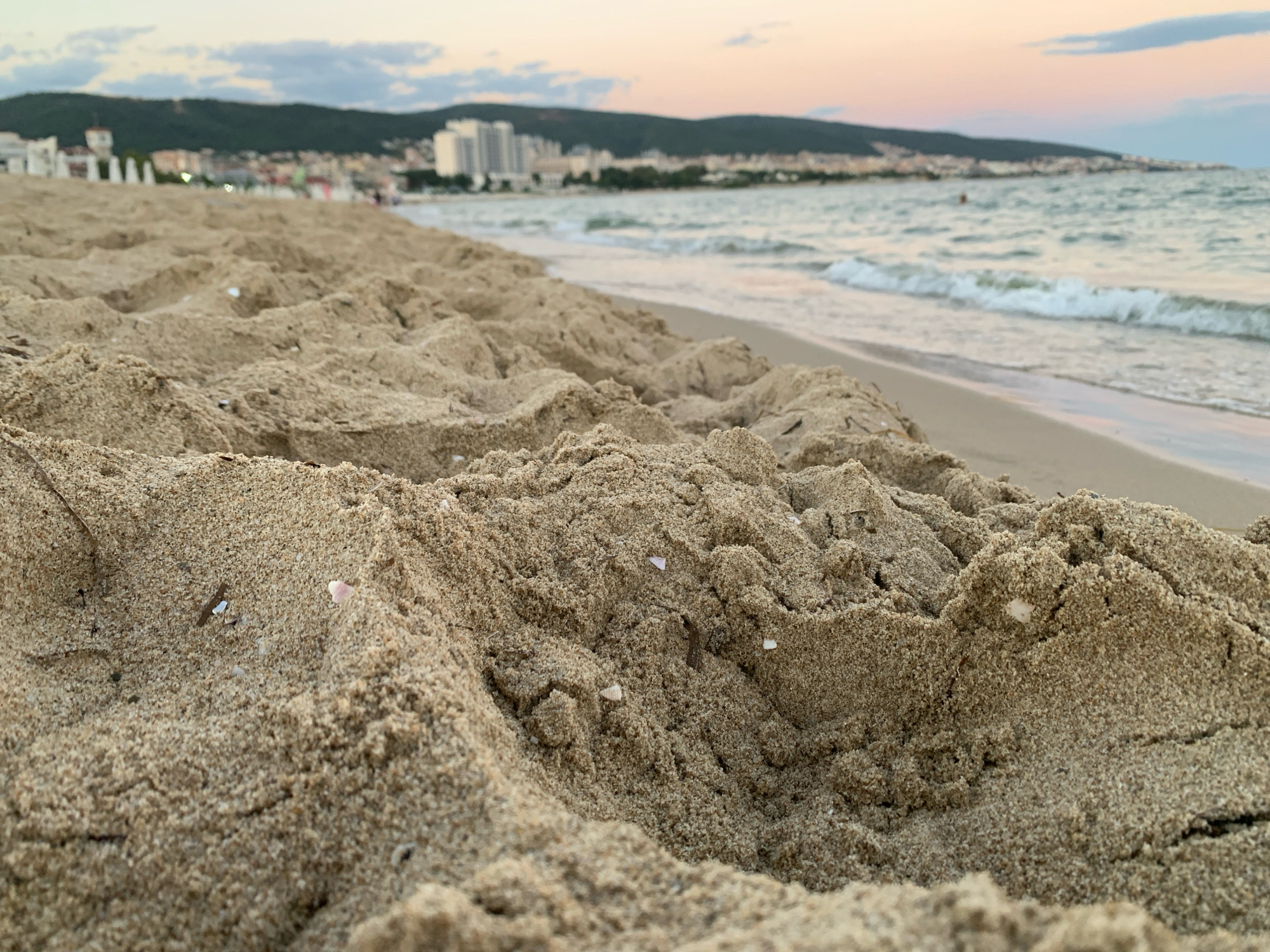 The image depicts a close-up view of sand dunes on a beach, with the sea and the city of Nesebar, Bulgaria, in the background. The foreground is dominated by textured sand formations, with small shells and debris scattered throughout. The middle ground features the gentle waves of the sea lapping at the shore. In the distance, the cityscape of Nesebar is visible, characterized by buildings and a hazy, overcast sky. The overall scene is peaceful and serene, conveying a sense of a quiet evening at the beach.