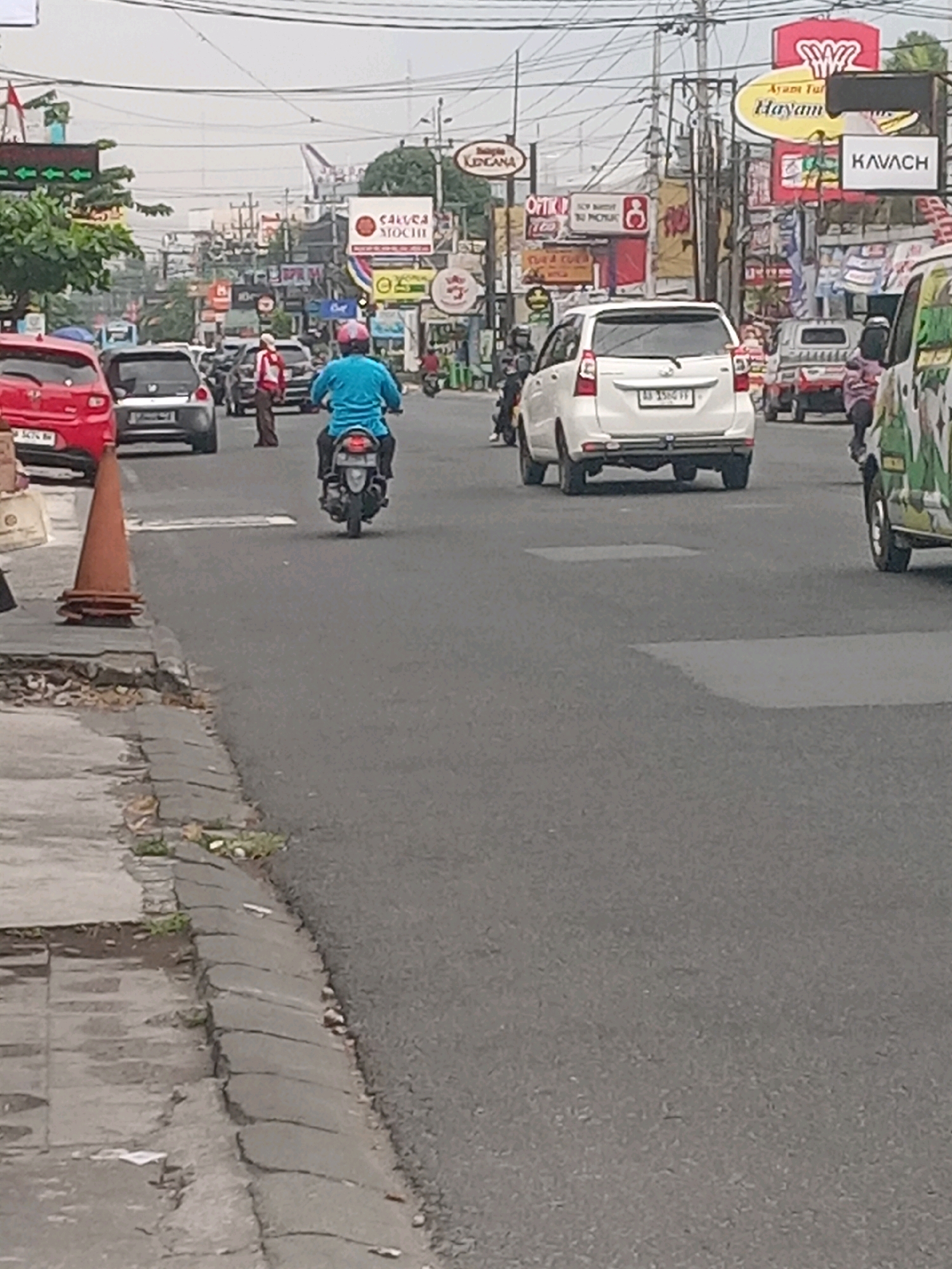 The image shows a street scene in Depok, Indonesia, on an overcast day. In the foreground, a traffic cone is placed on the sidewalk. The road is paved and has white lane markings. A person on a motorcycle wearing a blue shirt and a pink helmet is riding in the right lane. Several cars are also visible on the road, including a red car on the left, a white SUV, and other vehicles further down the street. Buildings with various signs line both sides of the road. One sign reads "AYAM TULANG HAYAM," and another says "KAVACH." The environment appears to be a commercial area with shops and businesses. The sky is cloudy, suggesting a lack of direct sunlight.