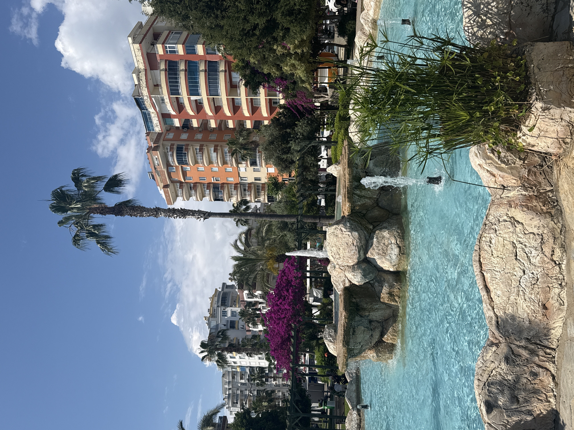 The image shows a waterfront scene in an urban environment. A multi-story building with orange and red accents stands tall in the background, partially obscured by trees. In the foreground, a man-made water feature is prominent, with rocks forming the edges of the pool. Water fountains spray upwards from the pool. Lush green plants grow near the water's edge. A tall palm tree is visible on the left side of the image, reaching towards a blue sky with scattered white clouds. Other buildings and greenery can be seen in the distance.