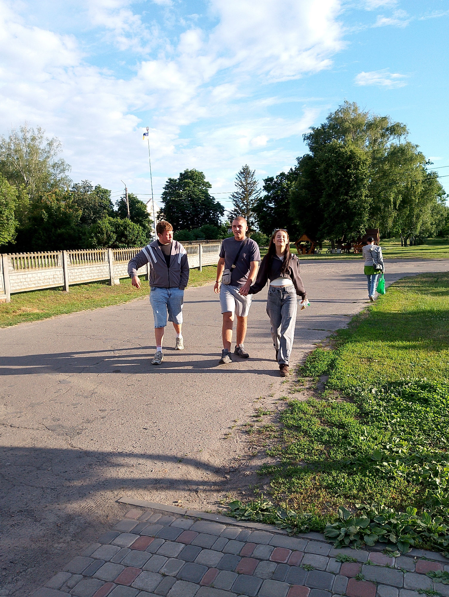 This image depicts a sunny outdoor scene in the village of Ustyvytsia, Ukraine, likely during the daytime. Three young adults are walking on a paved path. On the left, a young man wearing a grey hooded jacket and blue denim shorts walks with his hands in his pockets. In the center, a young man in a grey t-shirt and grey shorts walks, holding hands with a young woman on his right. The woman is wearing a dark cropped jacket over a light-colored top and grey pants, and she is smiling. Behind them, further down the path, a person wearing a grey jacket and green pants walks away from the camera, carrying a green bag. The path is bordered by a grassy area on the right and a fence with a brick wall and white pillars on the left. Trees and foliage are visible in the background, and a flagpole with a Ukrainian flag can be seen in the distance. The sky is blue with scattered white clouds. The overall atmosphere suggests a casual outing or stroll.