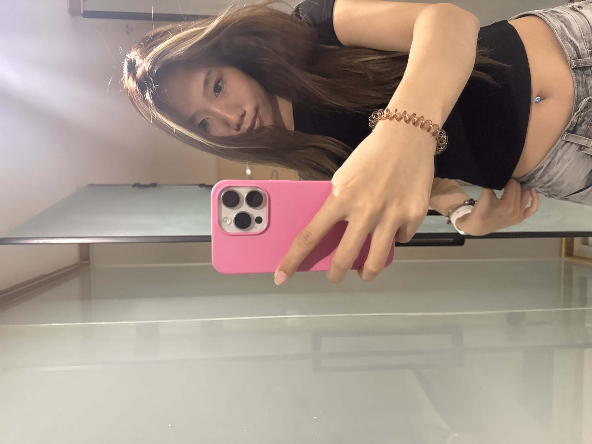 The image shows a mirror selfie of a young person, rotated 90 degrees clockwise, giving the impression that the subject is positioned on their left side. The individual has long, wavy brown hair with lighter highlights, cascading over their shoulder and partially obscuring the left side of their face. They look towards the camera with a neutral expression.
The person wears a black, short-sleeved crop top, revealing their midriff, which features a navel piercing with a prominent light blue gem. Light-colored denim shorts or jeans are visible from the waist down. Their right arm is extended, holding a bright pink smartphone, identifiable as an iPhone with a triple camera system. A translucent, brownish spiral-shaped bracelet adorns their right wrist. A white watch is also visible on their left wrist.
The setting is an indoor space, reflected in a large mirror. The background shows a light-colored wall or ceiling in the upper left, a distinct dark horizontal bar or shelf, and a lighter, possibly frosted glass or reflective surface directly below it. The scene suggests a casual moment, possibly taken in a dressing room or bathroom in Yuzhong District, China. There is no visible text in the image.