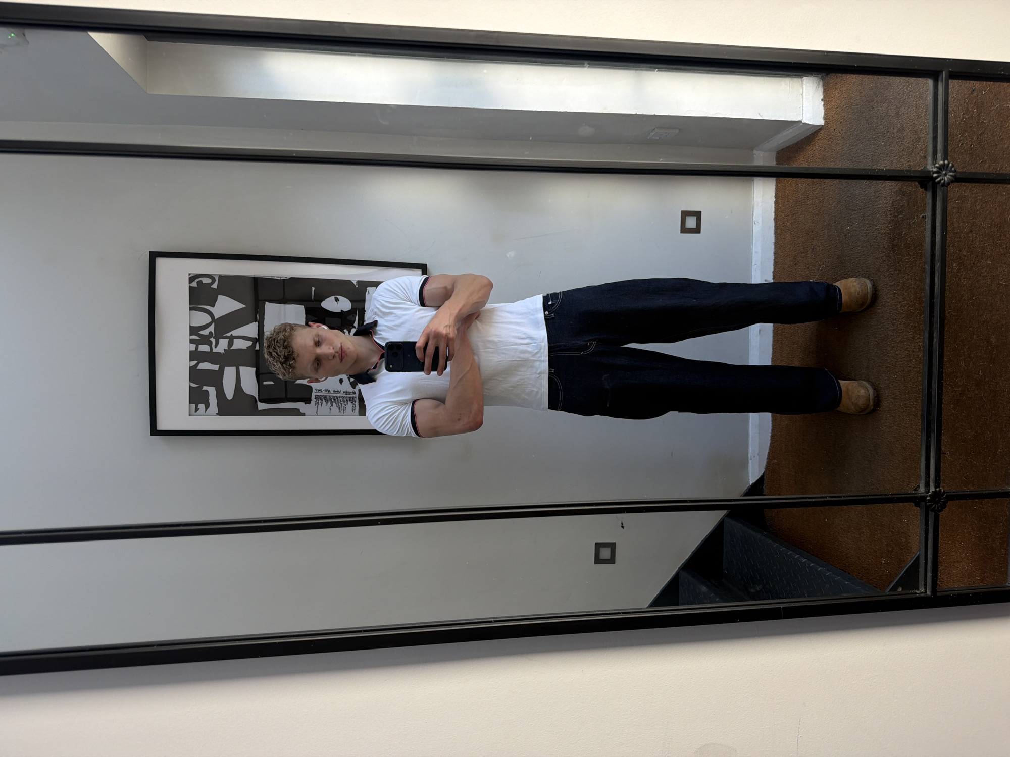 A young man with blond curly hair and white earbuds is taking a mirror selfie in what appears to be an indoor setting. He is wearing a white polo shirt with red and blue trim on the collar and sleeves, and dark wash, baggy jeans. He is holding a black smartphone in his right hand, capturing his reflection. The background shows a wall with a large framed black and white artwork, a tall mirror with a black frame, and a glimpse of a staircase. The floor is covered with a brown carpet. There is no visible indication of the location being in London, United Kingdom.