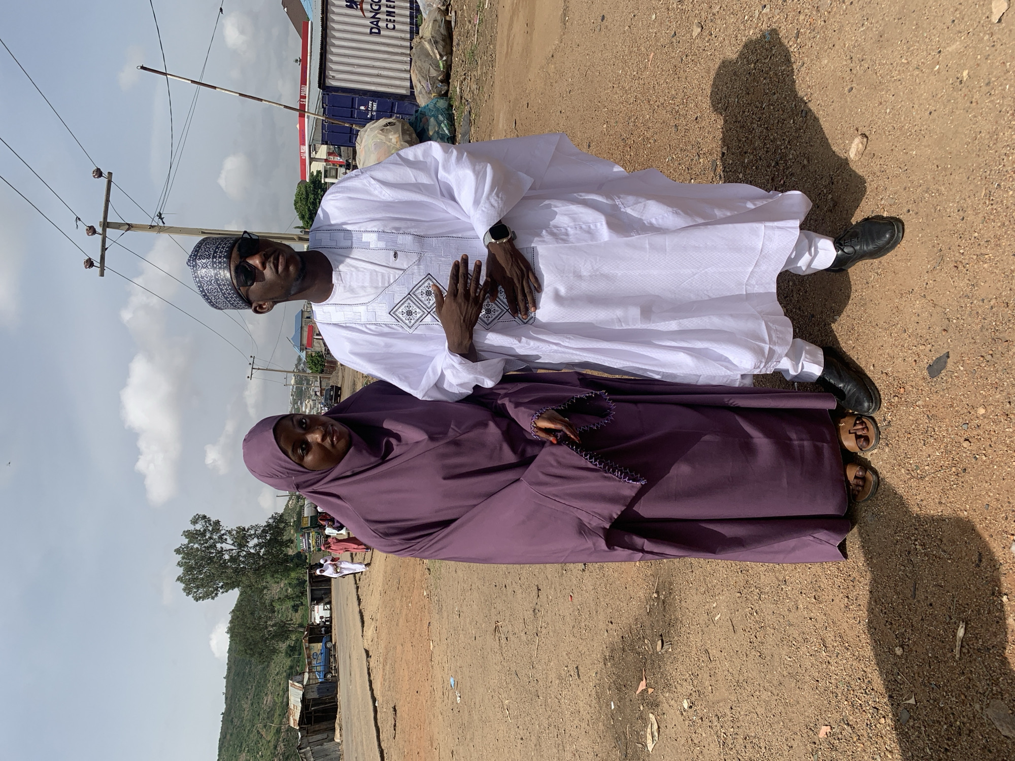 A man and a woman are standing in a dirt lot in Naraguta Hausawa, Nigeria. The man is wearing a white traditional Nigerian outfit with black shoes, sunglasses, and a patterned cap. He is wearing a watch on his left wrist. The woman is wearing a purple hijab and holding prayer beads. A mountain is in the background, with small buildings scattered around the area. Several utility poles and electrical wires are in the background, and some trucks and shipping containers are visible. A shadow is visible of the man. The lighting is bright and the weather appears sunny.