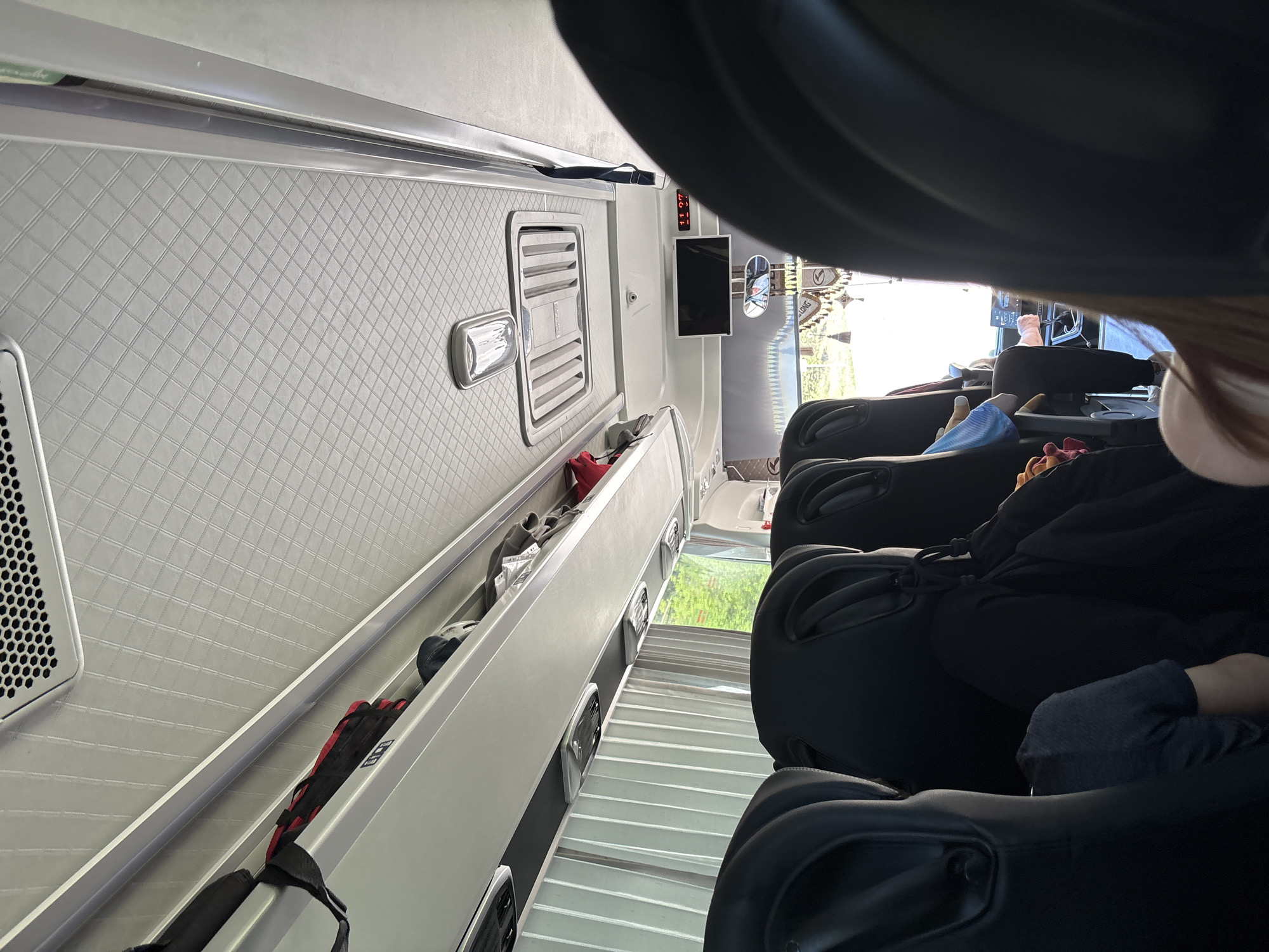 The image shows the interior of a luxury van or bus, possibly in transit. It features rows of reclining leather seats with passengers. Overhead, there are storage compartments with various items. There is a small screen displaying the time. The interior walls are light grey with a quilted pattern. The scene appears to be a bright day, as seen through the windows. There is no information about the location in the city of Votkinsk, Russia.