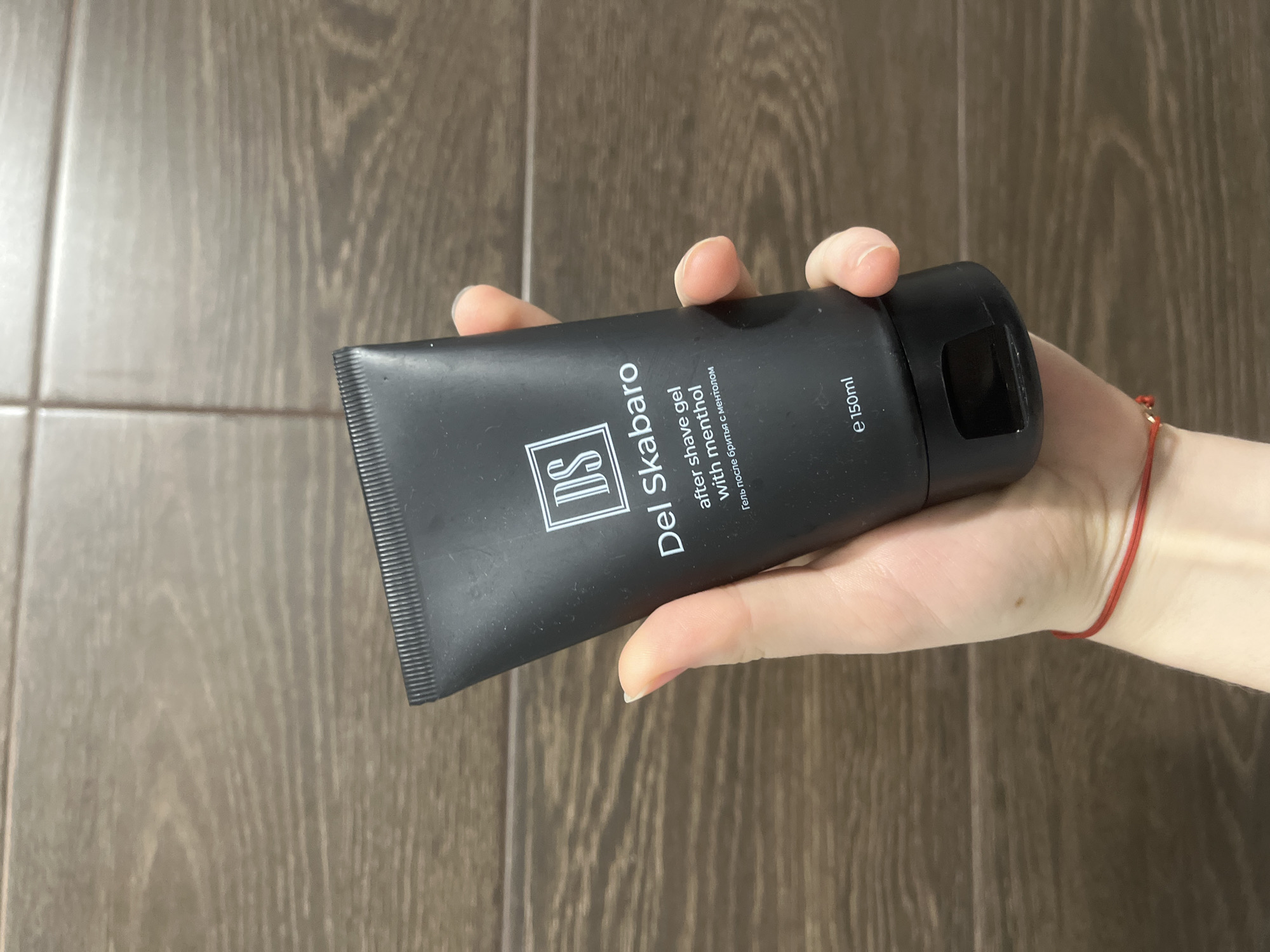 The image depicts a close-up scene located in the city of Saint Petersburg, Russia, featuring a person's hand holding a matte black cosmetic tube against a tiled background. The tube is a "Del Skabaro after-shave gel with menthol," as indicated by the white text on its surface, which also includes the Russian translation "Гель после бритья с ментолом" and "e 150ml." A white "DS" logo in a square is visible near the top of the tube. The hand holding the tube has light skin, with visible fingers (thumb, index, middle, ring, pinky) and short, naturally colored nails. A thin red string bracelet is present on the wrist, along with a small, light brown mole on the back of the hand. The background consists of brown-toned rectangular tiles with a wood grain texture, arranged with distinct grout lines, suggesting an indoor bathroom or similar setting. The lighting is even.