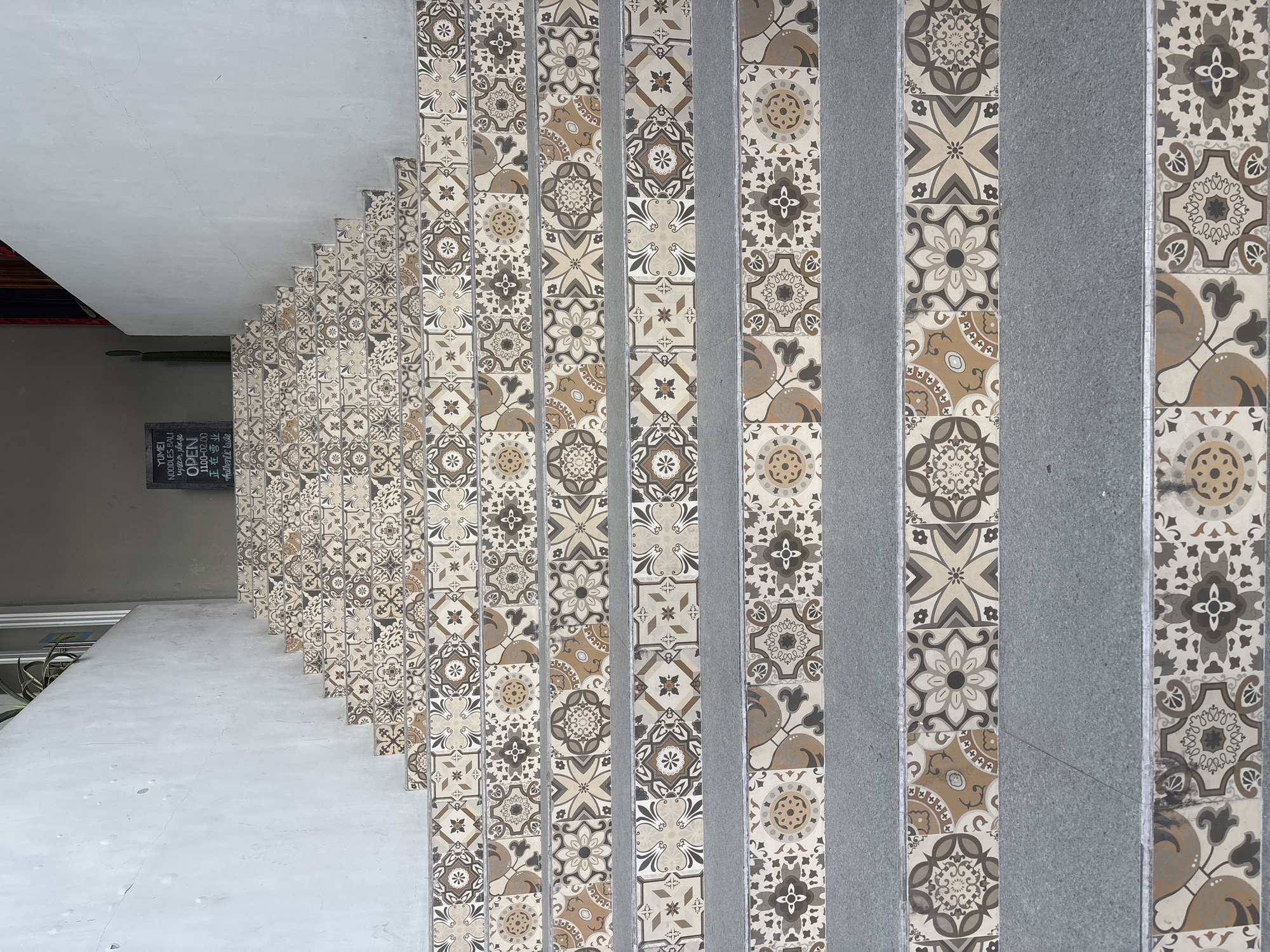 The image shows a staircase in North Kuta, Indonesia. The steps are tiled with a patterned design in shades of beige, brown, and gray. A gray panel runs alongside the tiled section of each step. A white ceiling is visible above the staircase. A chalkboard sign is on the wall, advertising "Yumei Noodles Bali", with the text "upstairs please", alongside the hours of operation and the words "Authentic taste".