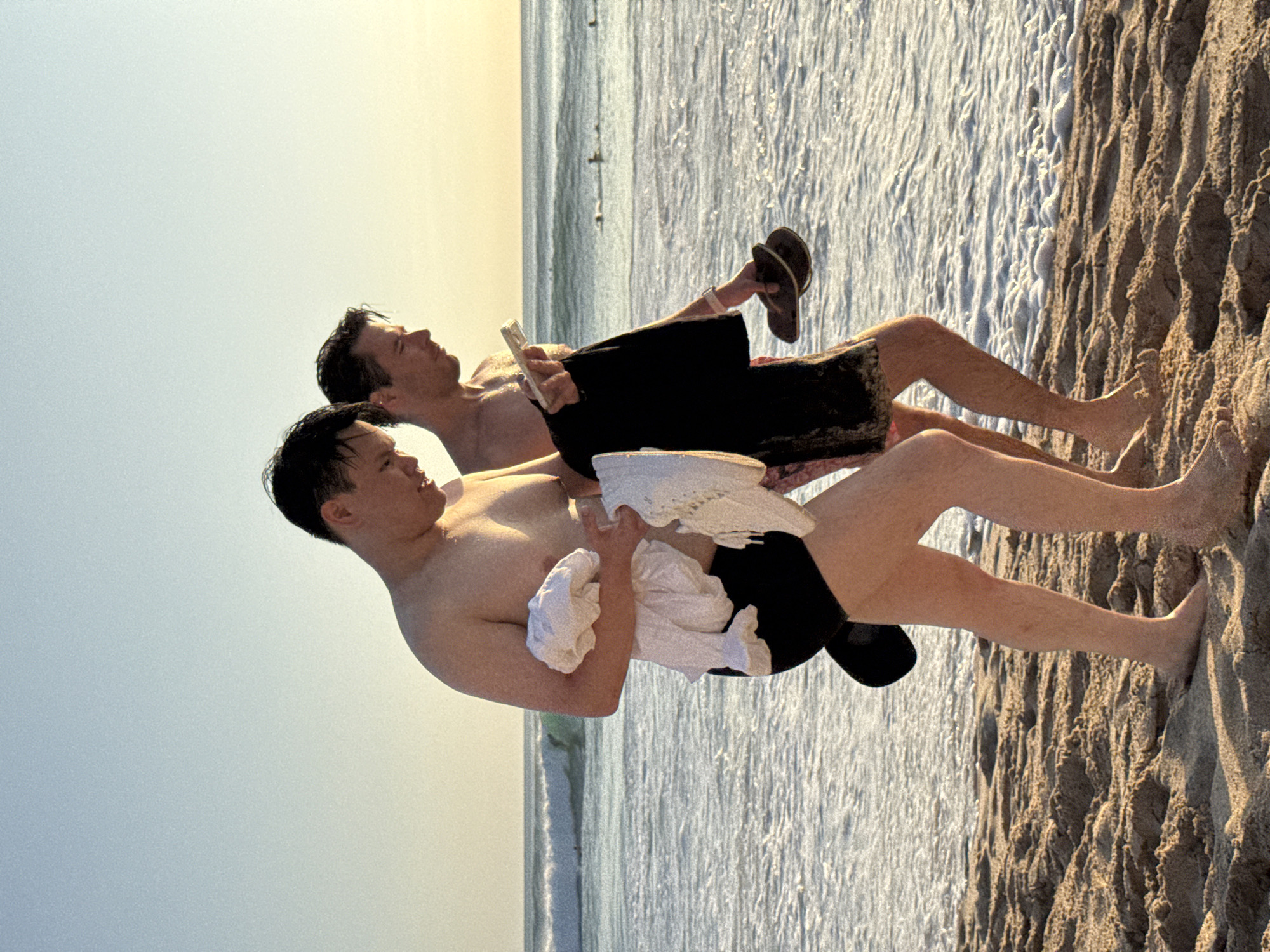 Two shirtless men are reclining on a sandy beach next to the ocean. The man in the foreground is holding a white towel and wearing black shorts. The man in the background is wearing black shorts and holding a book. One hand holds a pair of sandals. The sand is a dark brown. The ocean is light blue and the sky above is lighter. This image could be taken in Kuta Selatan, Indonesia.