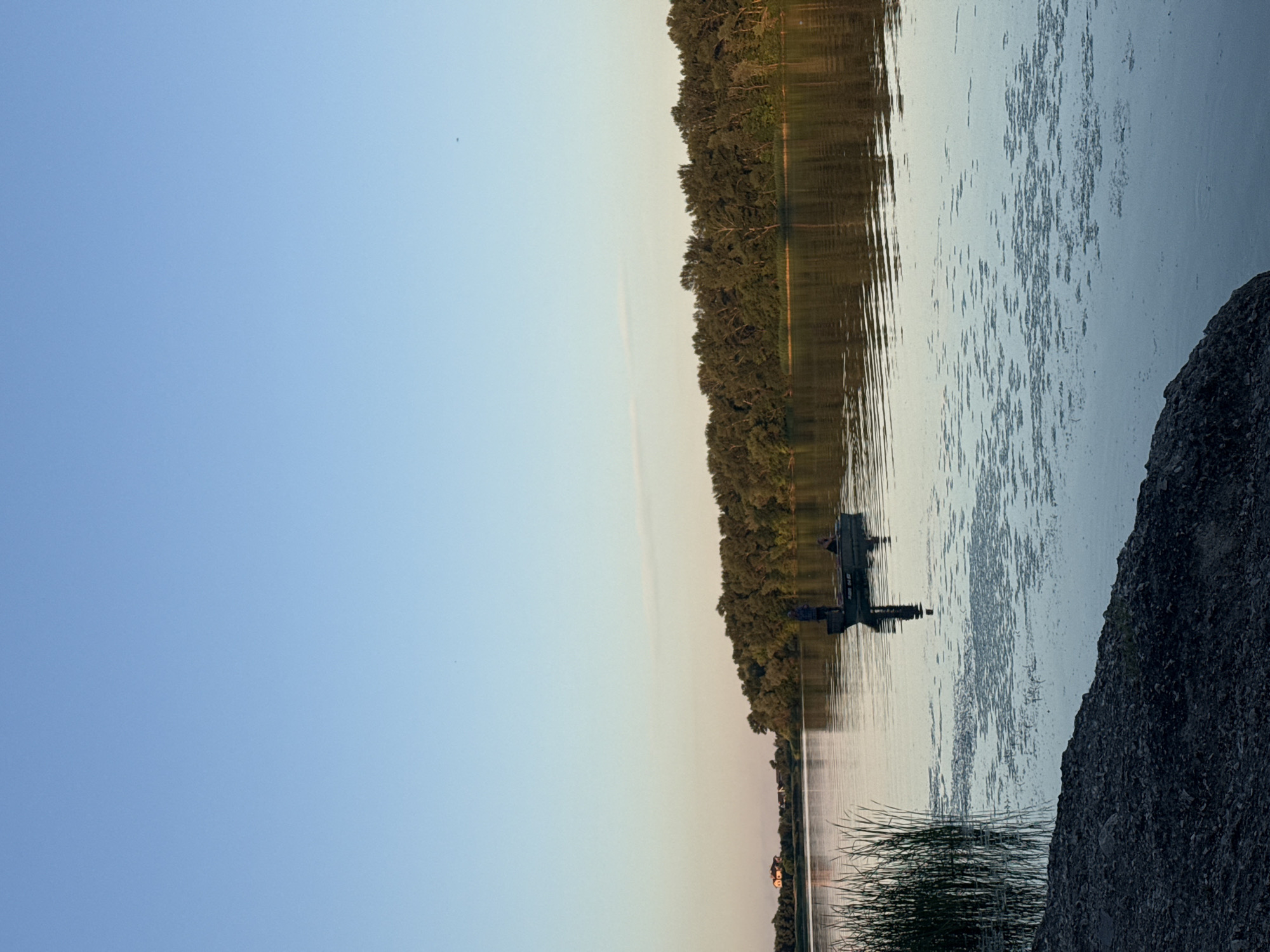 A body of water, likely a river, is depicted with a distant tree line forming the horizon. In the foreground, rough, dark-colored rocks line the bank of the water. Tall reeds grow from the water's edge. A small, dark-colored motorboat is in the middle ground of the water. Two individuals are in the boat; one is standing and appears to be fishing. The water's surface has small patches of aquatic vegetation. The sky above is a clear, pale blue, suggesting either early morning or late evening light. The scene is peaceful and serene, with the reflections of the trees and sky visible on the calm water. It's not possible to determine the specific location within the city of Dnipro, Ukraine from the visual information provided.