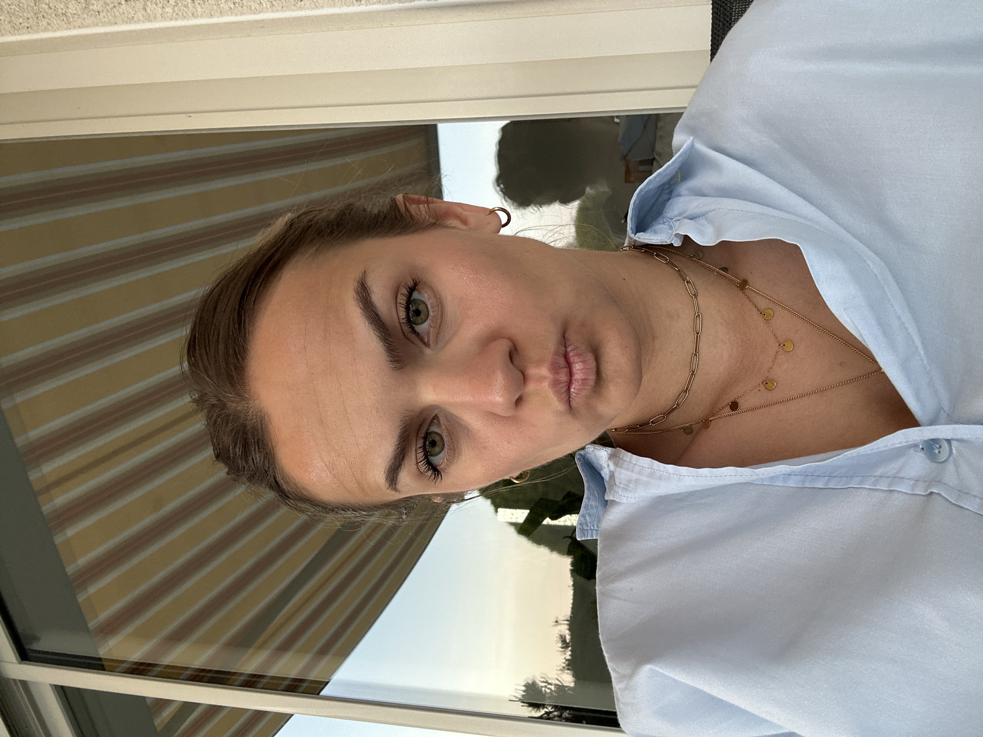 A woman is facing the camera, likely indoors near a window. Her expression is neutral. She is wearing a light blue shirt, with two gold necklaces visible on her neck. The window has striped blinds. The background shows a hint of greenery outside. The location is Unknown, Unknown.
