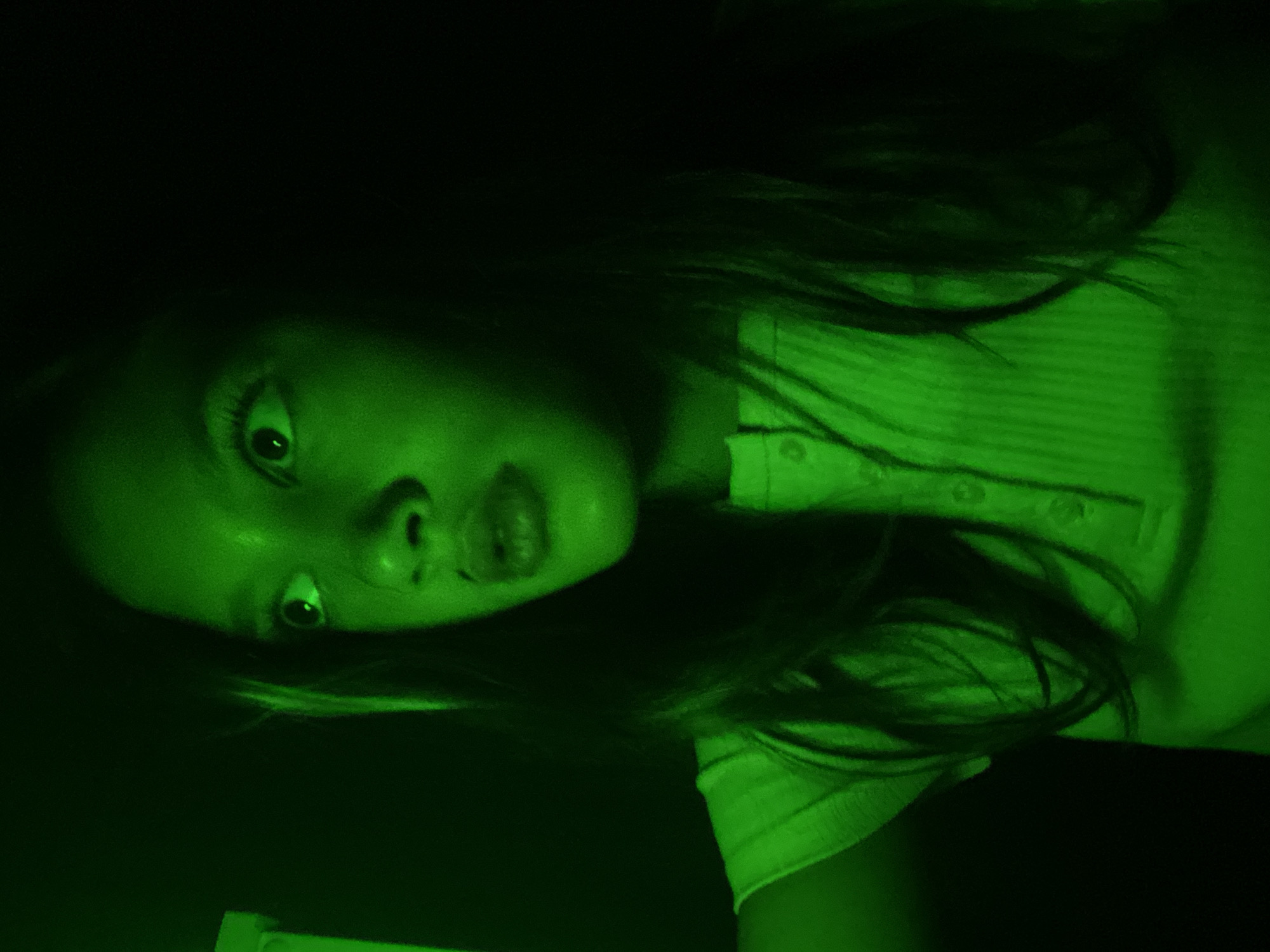 The image is a close-up, over-the-shoulder shot of a young female looking upwards and to the left with a surprised or concerned expression. She has long, dark hair and is wearing a light-colored, collared shirt with buttons. The scene is lit with a strong green light, obscuring many details of the background, which appears dark. The girl's eyes are wide, and her mouth is slightly open. The image quality is grainy and somewhat blurry. There is no indication of the specific location within Novosibirsk, Russia, as the image provides no recognizable landmarks or context.