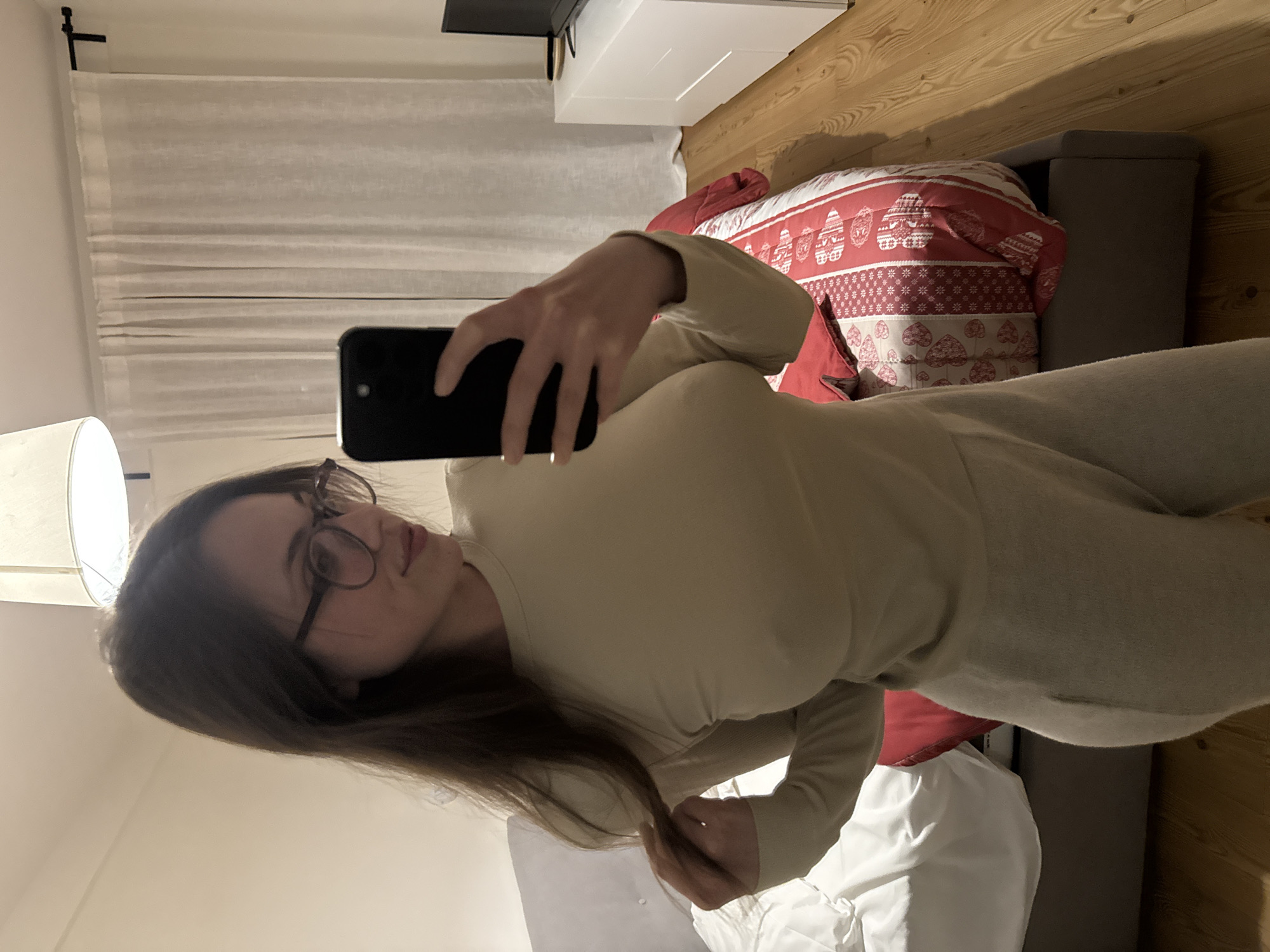 A woman with brown hair and glasses is taking a selfie in a bedroom. She is wearing a beige top and matching pants. In the background, there is a bed with red and white bedding, a wooden floor, white curtains, and a white cabinet. The location is likely Sappada / Plodn / Sapade, Italy, as the user specified it in the prompt.