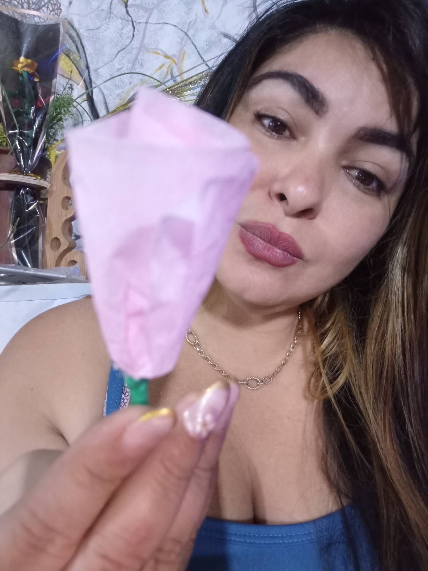 An adult female is depicted in a close-up indoor portrait, originating from Juan Griego, Venezuela. She is shown from the upper chest upwards, looking directly towards the viewer with her head slightly tilted. Her right hand is extended into the foreground, holding a light pink, crinkled object that resembles a stylized or artificial flower bud. The visible fingernails on her right hand include one with gold polish on the tip and others with a light, possibly iridescent, polish.
The woman has brown eyes, defined dark eyebrows, and her lips are colored with a reddish-pink lipstick. She wears a gold-colored chain necklace around her neck and a blue garment, a strap of which is visible over her right shoulder. Her long, dark brown hair with lighter highlights is visible cascading over her right shoulder.
The background is softly blurred, revealing a light-colored wall with abstract dark and gray lines. Partially discernible decorative elements in the background include what appears to be a yellow flower wrapped in clear plastic, some green foliage, and a wooden object with rounded cutouts. The overall lighting suggests an indoor setting.