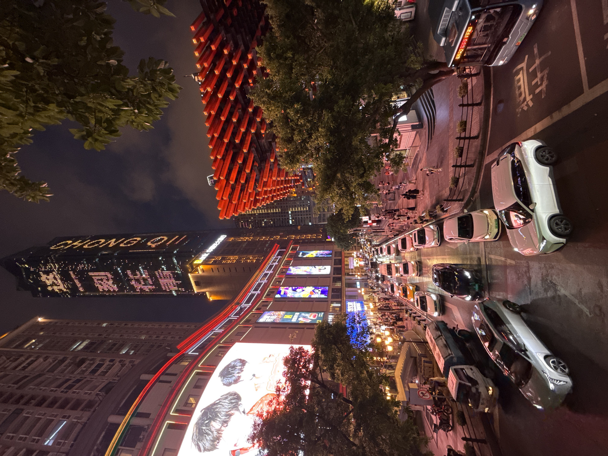 This is an eye-level, full-frame, wide shot taken at night in the Yuzhong District of China, likely Chongqing. The scene depicts a bustling city street at night, illuminated by artificial lights from buildings and street lamps. A tall skyscraper in the background displays illuminated text that reads "CHONGQING" vertically. In the foreground, a multi-lane road is congested with traffic, including a silver SUV, a white SUV, a dark sedan, a yellow taxi, and several other vehicles. A large white truck is also visible. Pedestrians are walking on the sidewalks and crossing the street. To the right, a red, lattice-like structure is prominently featured, with trees in front of it. A blue bus is also present in the lower right corner of the image, with Chinese characters on its side. The overall impression is of a vibrant urban environment at night.