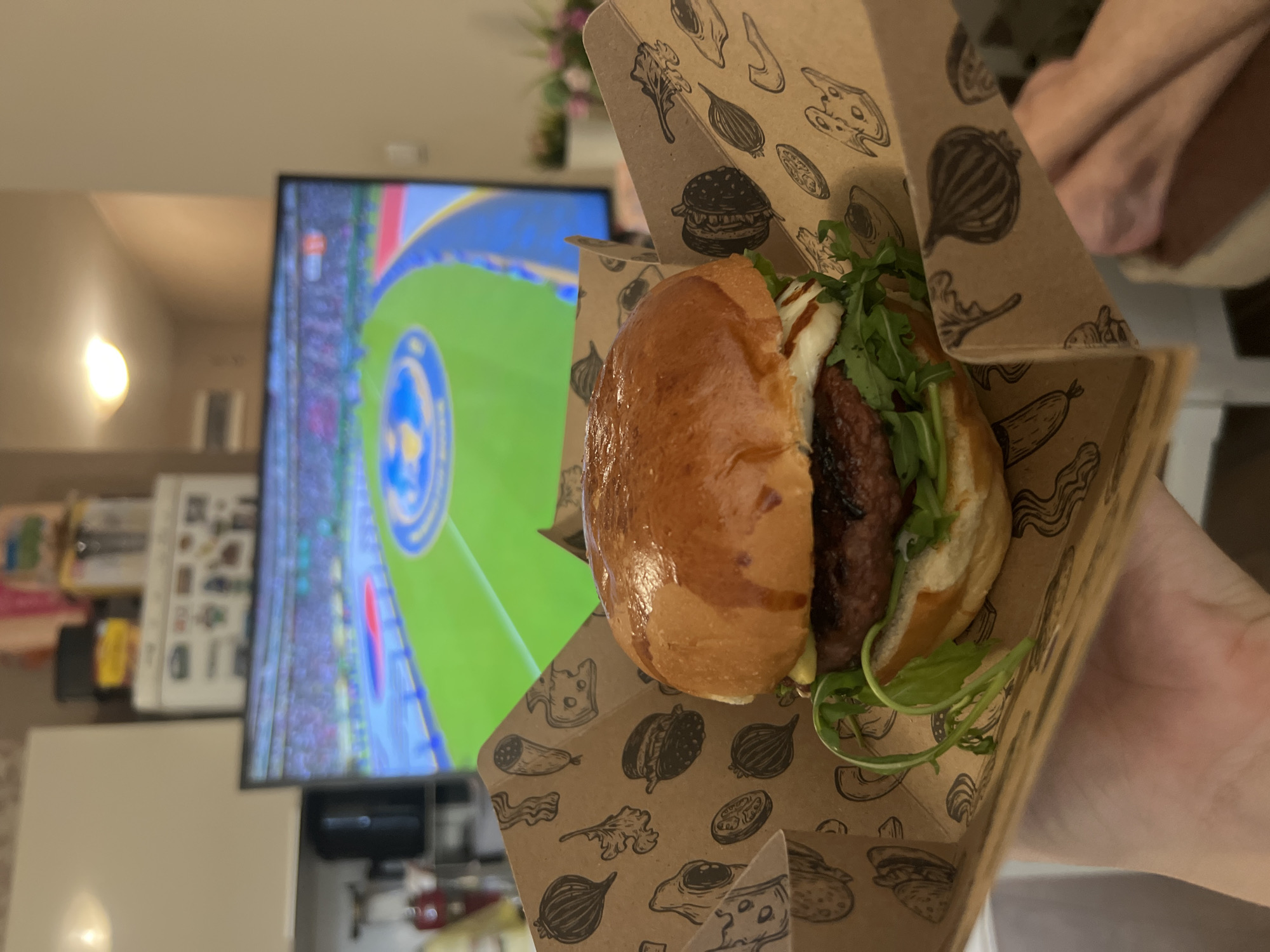 A close-up shot shows a hand holding a burger in a brown cardboard container. The burger is on a bun and topped with lettuce, cheese, and sauce. In the background, a television screen displays a sports game. The scene appears to be indoors, likely in a home or restaurant. The location is not identifiable as Sofia, Bulgaria, or any specific city.