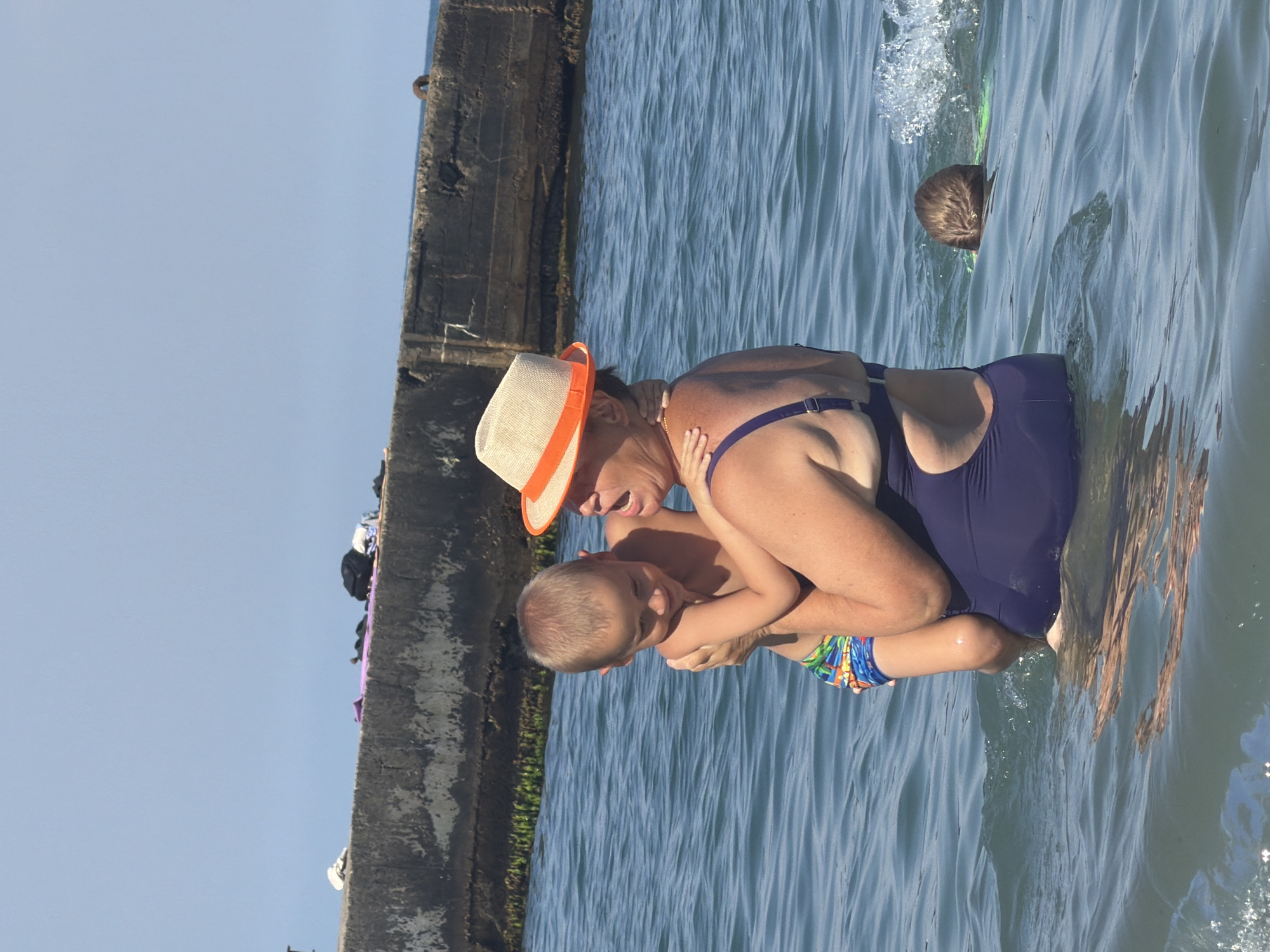 In Partenit, Ukraine, a woman wearing a navy blue swimsuit and a straw hat with an orange band holds a young boy in her arms while standing in the sea. The boy, wearing colorful swim trunks, is being held close to the woman's chest. In the background, another child with a light brown buzz cut is swimming in the water, with a splash of water visible near their head. A concrete structure, possibly a pier or breakwater, extends into the water behind the people, with some debris scattered on top of it. The sea is choppy, with visible waves. The sky is clear and blue.