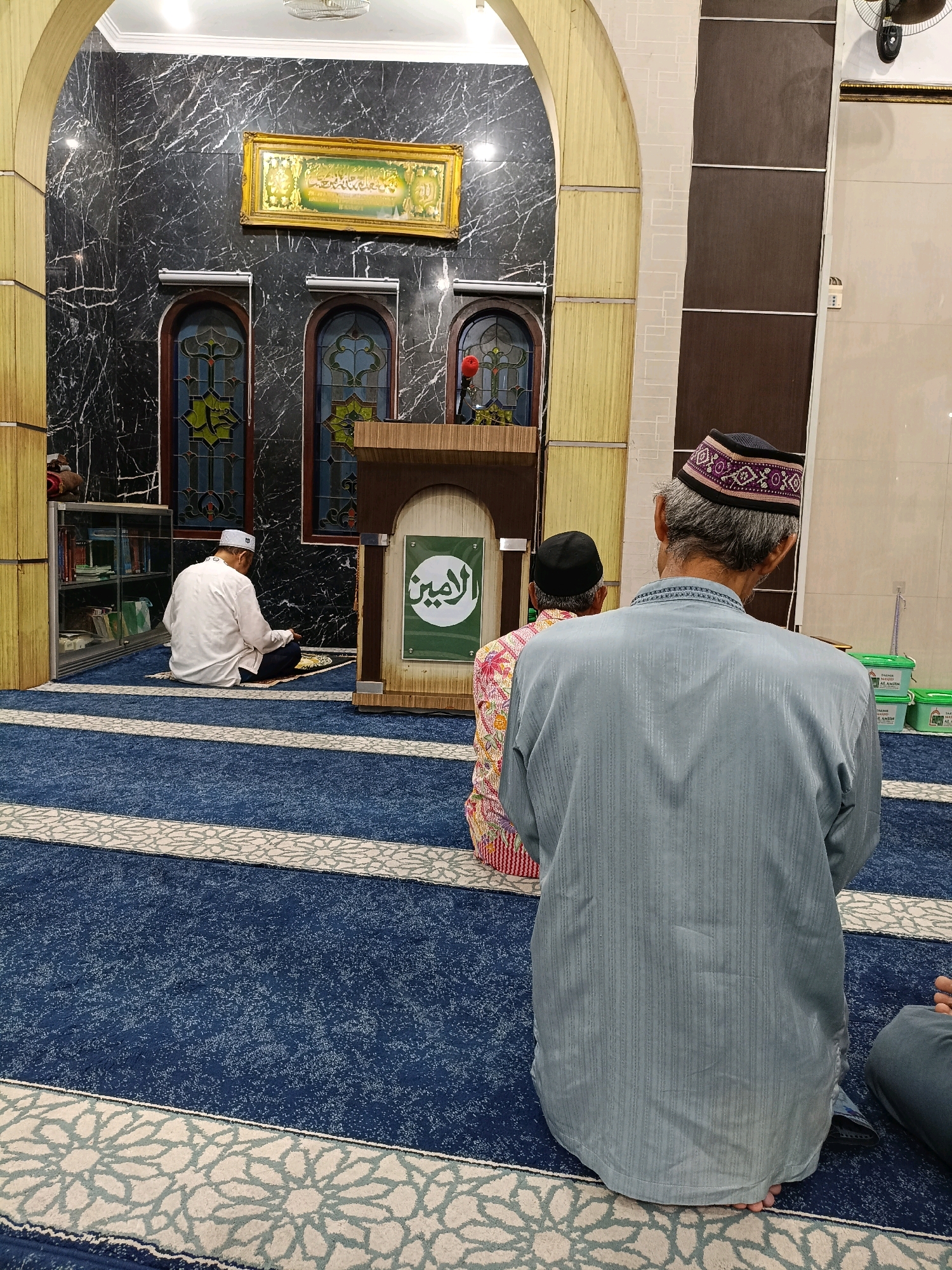 In Maguwoharjo, Indonesia, a mosque interior is depicted, featuring a blue carpet with a floral pattern and white borders. Three men are present, engaged in prayer. One man, wearing a white cap and shirt, sits on a prayer mat on the left. To his right, another man in a patterned shirt and black cap faces away from the camera. A third man, in the foreground and facing away, wears a grey tunic and a patterned cap. Behind the man in the grey tunic, a wooden podium with Arabic calligraphy on a green background is visible. The mosque walls are adorned with black marble and decorative arched windows. A framed artwork hangs on the wall. The scene conveys a sense of religious devotion within a traditional mosque setting.