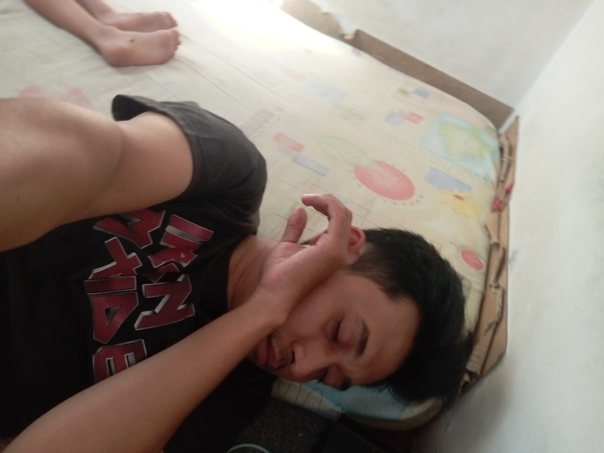 The image depicts a man sleeping on his side on a mattress with a patterned sheet. His eyes are closed, and his right hand is raised to his face, covering his mouth and nose. He is wearing a black t-shirt with red lettering. In the background, another person's legs are visible, also appearing to be lying down. The setting appears to be indoors, possibly a bedroom, with a white wall visible to the right. The lighting suggests it is daytime, with natural light streaming in from the left. The overall impression is a casual, relaxed scene of people resting.