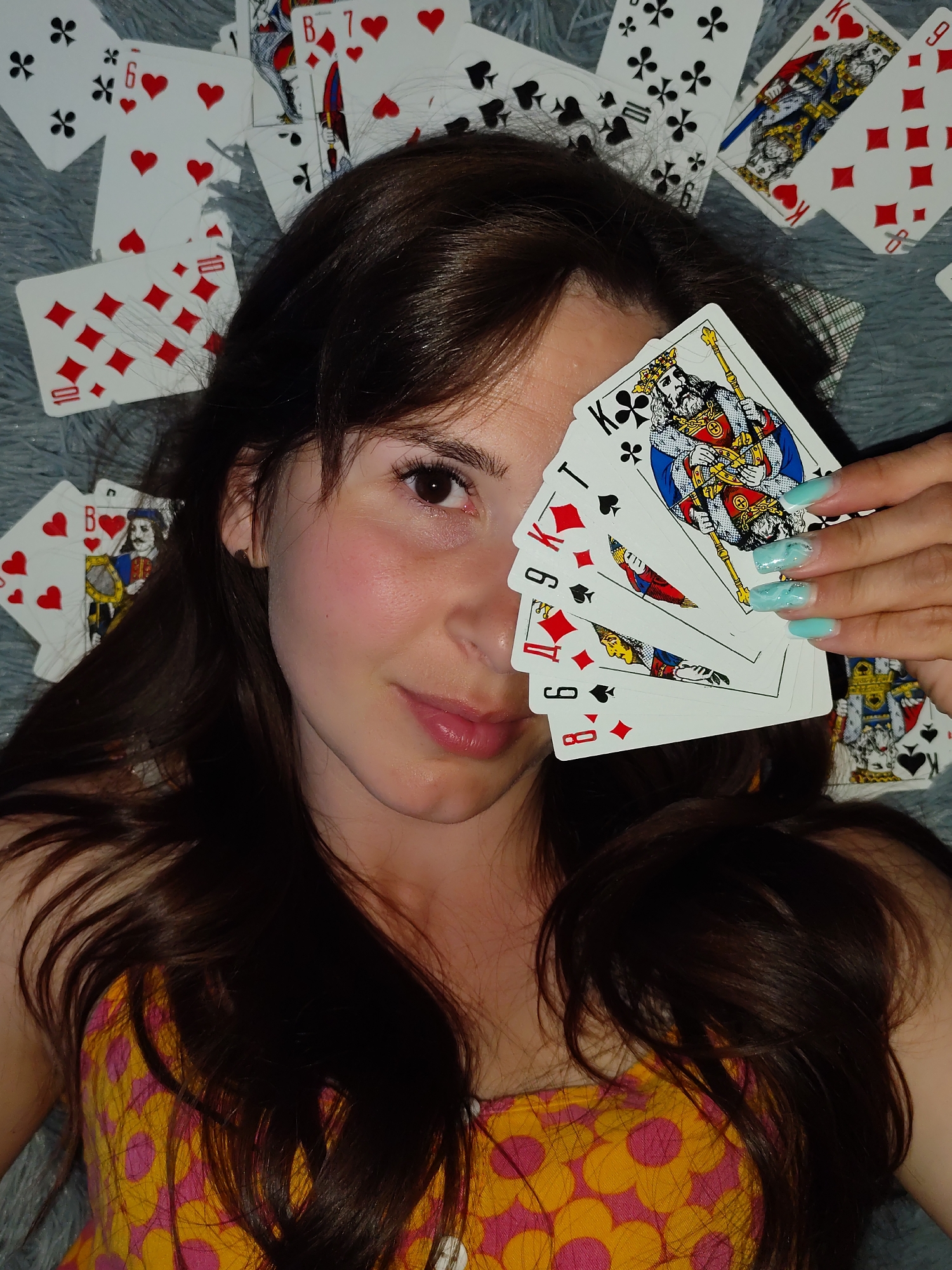 A fair-skinned woman with long, dark hair is the focal point, holding a hand of playing cards partially obscuring her face. The cards include a King of Clubs, Ten of Spades, Nine of Diamonds, Nine of Spades, and Eight of Diamonds. She has long, light blue fingernails with white swirls. The cards are against a background of more playing cards scattered on a grey, textured surface, possibly a blanket or rug. She wears a yellow and pink floral top. The location is not discernible from the image and could not be identified as Фоки, Russia.