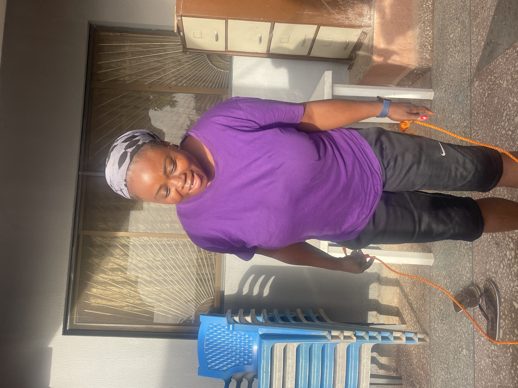 This image captures a joyful woman in a sunny, semi-outdoor setting in Jos, Nigeria. The central subject, a woman with dark skin, is smiling broadly at the camera, conveying happiness. She is wearing a vibrant purple short-sleeved t-shirt, black athletic shorts with a white "Nike" logo on the left leg, and a grey and black patterned headscarf. A blue wristband or watch is visible on her left arm, and her fingernails are painted red.
She is holding an orange rope with both hands, extending across the lower part of her body. The ground is a light-colored, speckled tiled surface, characteristic of an outdoor patio or covered walkway. Bright sunlight casts clear shadows, indicating it is likely mid-morning or afternoon.
In the background to the left, several blue plastic chairs are stacked neatly. To the right, a multi-drawer metal filing cabinet in a light brown or beige color is visible. A window with horizontal security bars or grills can be seen behind her. A pair of dark flip-flops rests on the ground near her right foot. The scene suggests a relaxed, casual environment, possibly a residential or communal area.