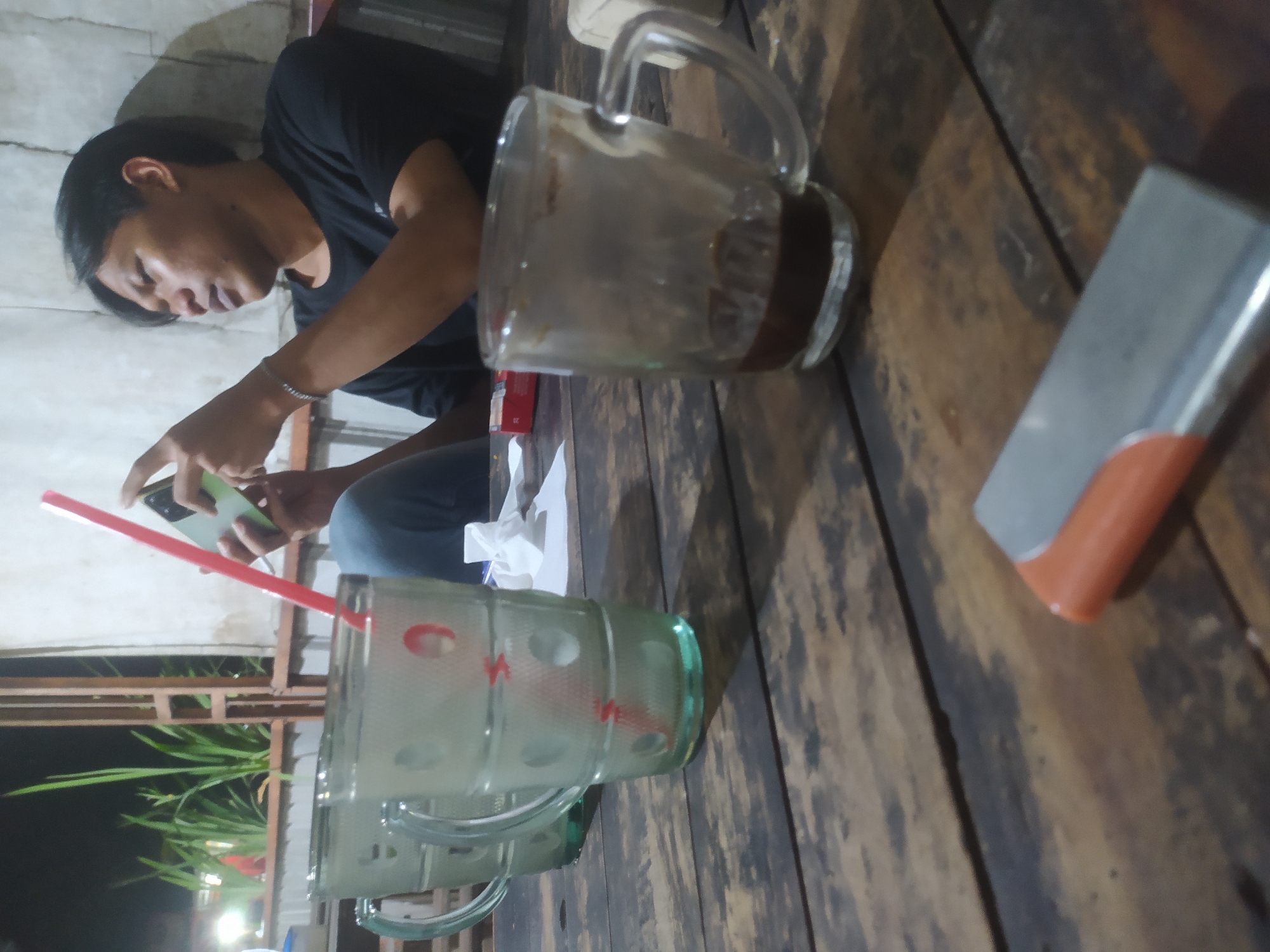 The image depicts a dimly lit, outdoor setting, possibly a roadside cafe or warung, in Tuntang, Indonesia. A young man with short black hair is seated at a rustic wooden table, engrossed in his mobile phone, which he holds in his right hand. He is wearing a black t-shirt. A red plastic straw extends from a glass on the table, near his phone. To his left, two more glasses, one of which appears to be filled with a light-colored beverage and also contains a red straw, are visible. In the foreground, a lighter with an orange and silver casing rests on the table. The background features some greenery and a concrete wall. The overall ambiance suggests a casual evening gathering.